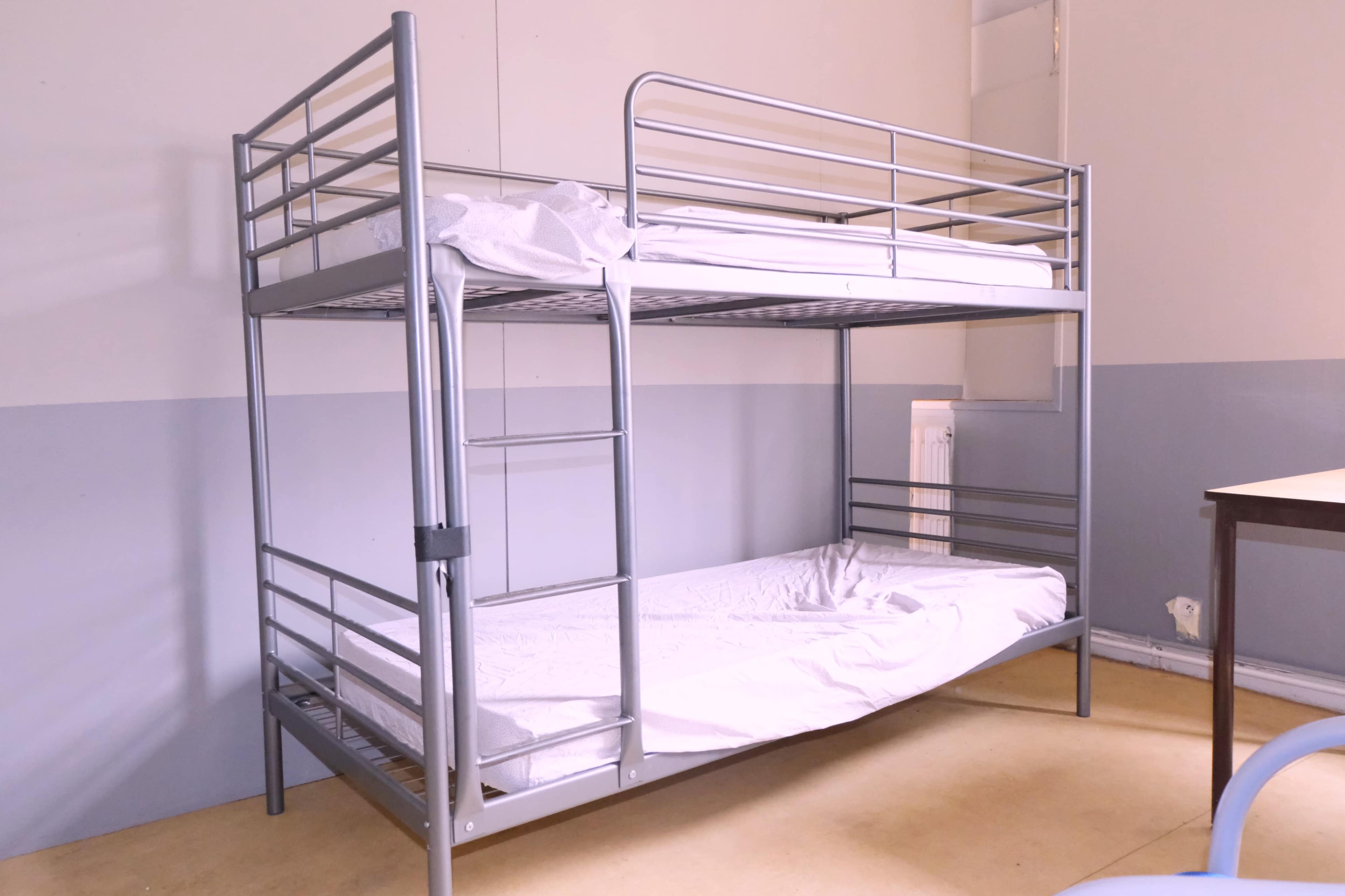 A gray metal bunk bed with two mattresses, one on top and one on the bottom, in a room with plain walls and minimal furniture.