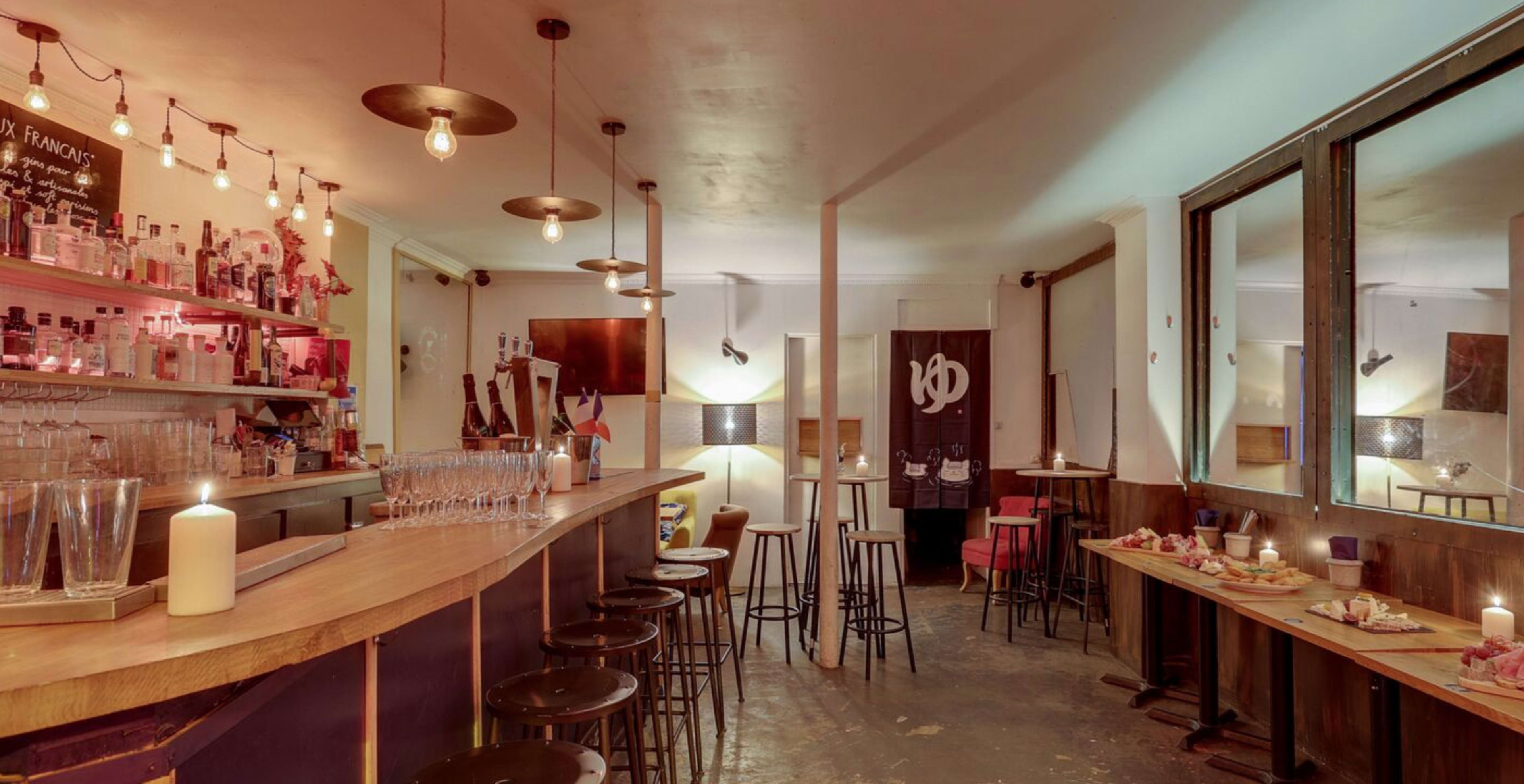 A dimly lit bar with a wooden counter, high stools, and a shelf of various alcoholic beverages.