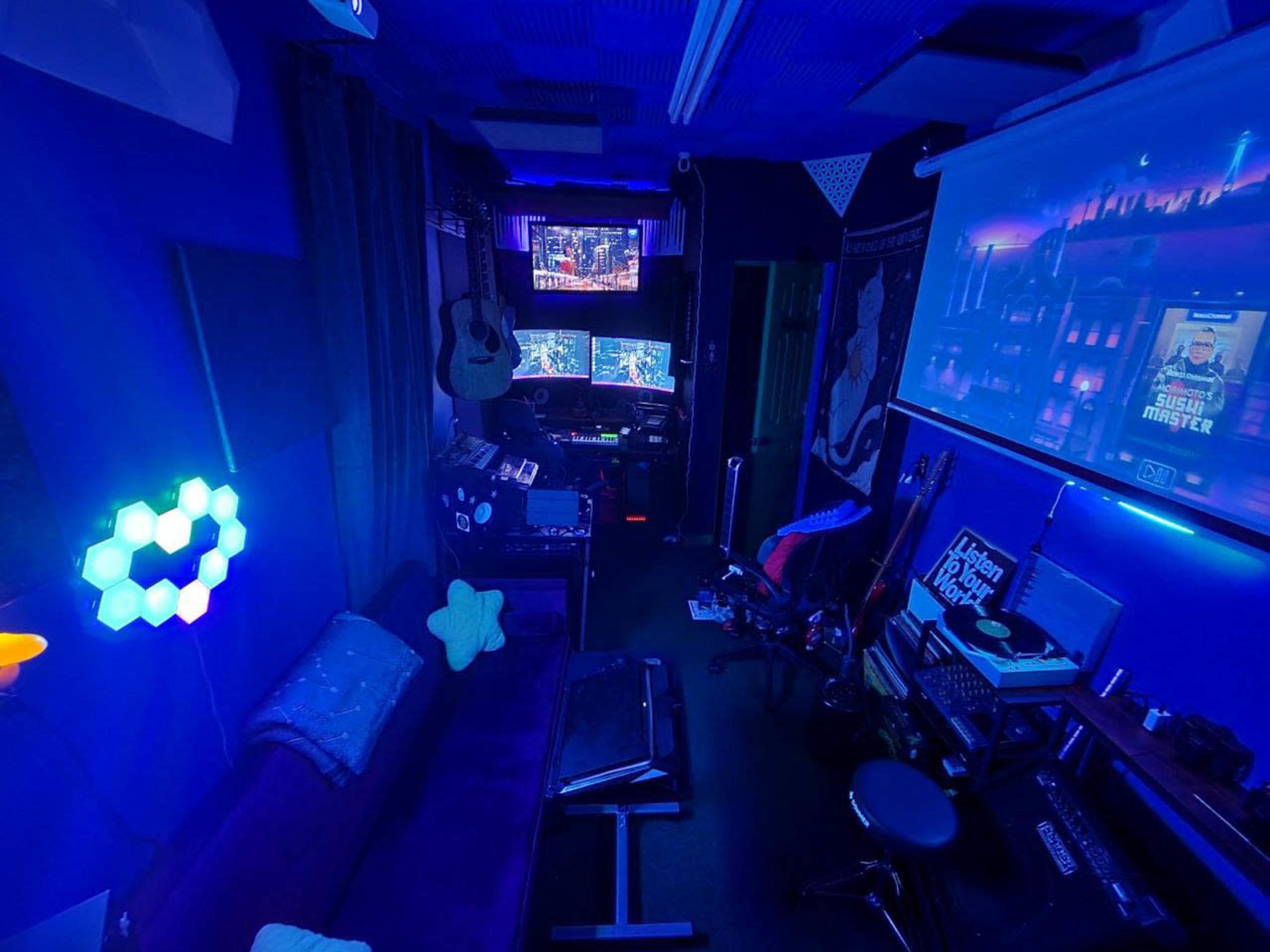 The image shows a dimly lit room furnished with musical instruments, multiple screens displaying graphics, and colorful LED lights.