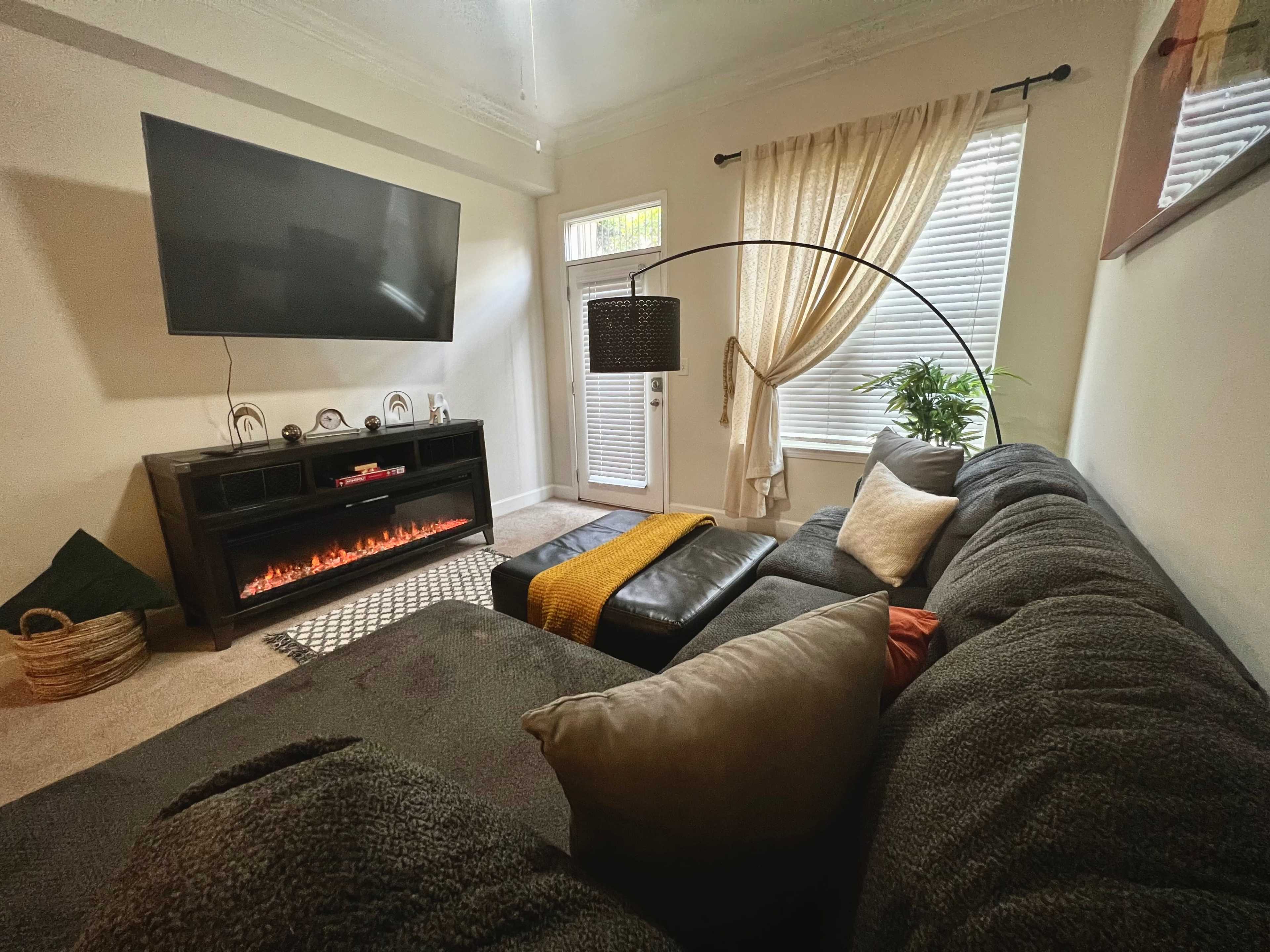 A sectional sofa is positioned in a living room with a wall-mounted TV and an electric fireplace beneath it, alongside large windows covered with light curtains.