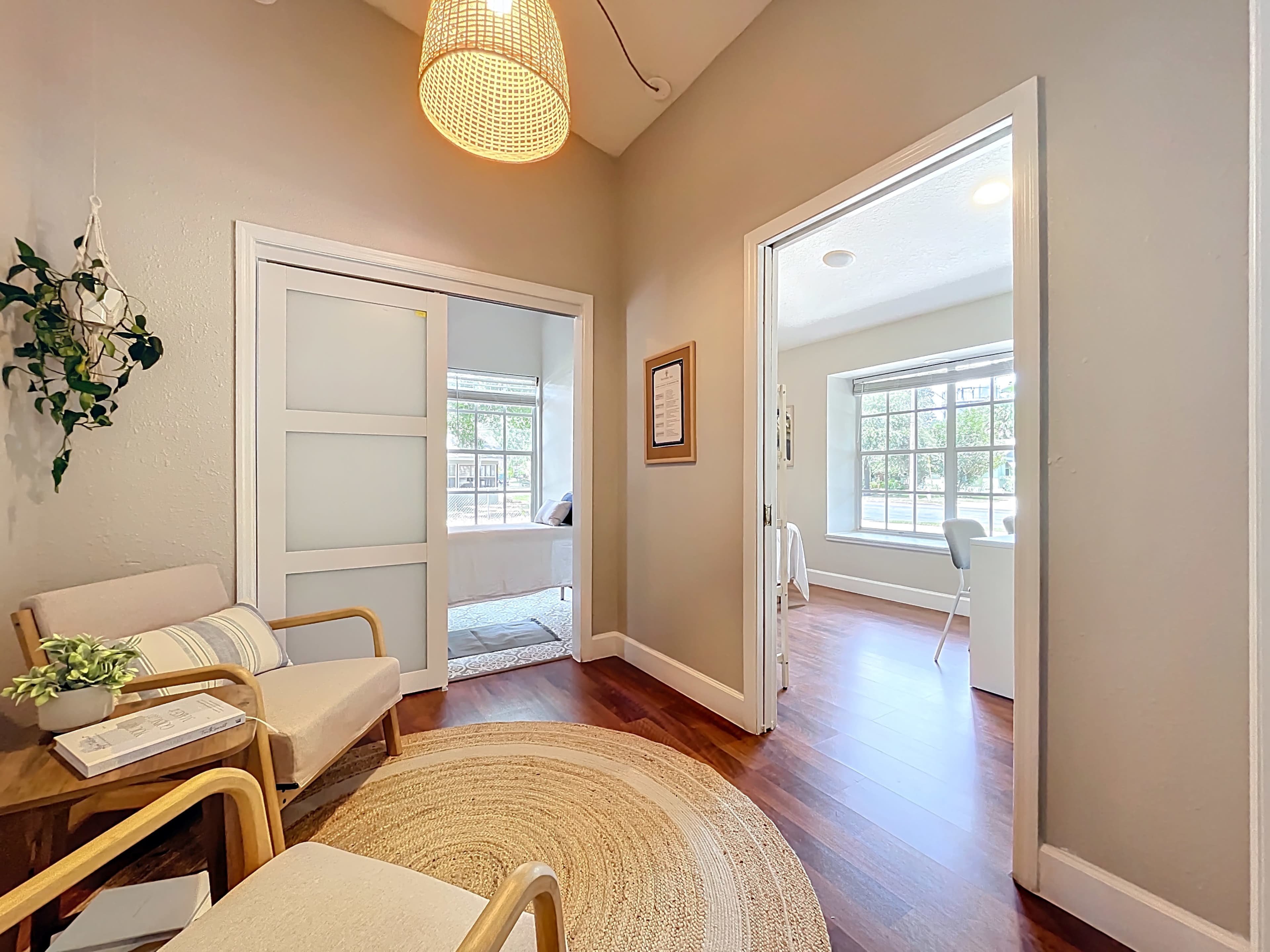 A bright entryway features a light fixture, a round rug, and two chairs, leading to rooms with large windows.
