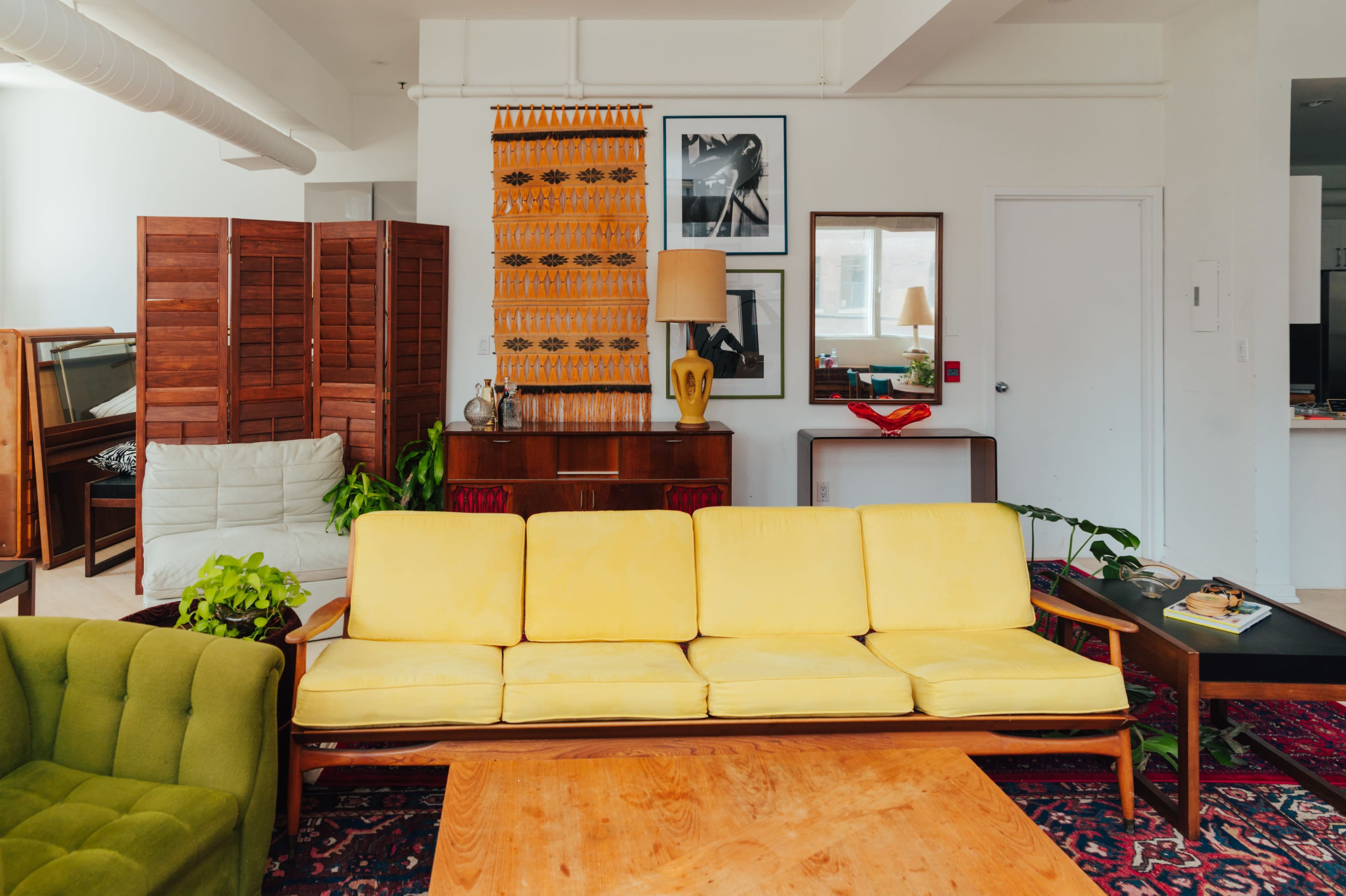 A brightly lit living room features a yellow sofa, a green chair, wooden furniture, and framed artwork on the walls, with plants adding touches of greenery.