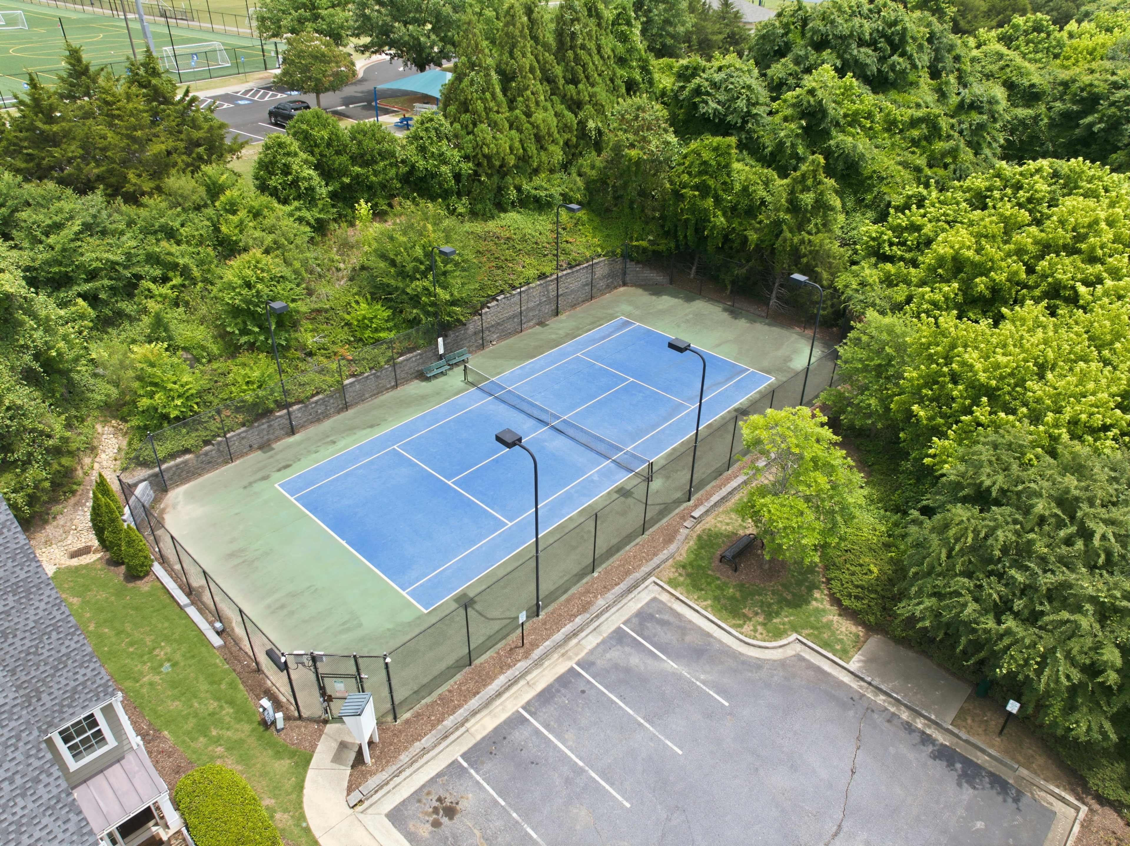 The image shows an aerial view of a tennis court surrounded by trees, with a parking area adjacent to it.