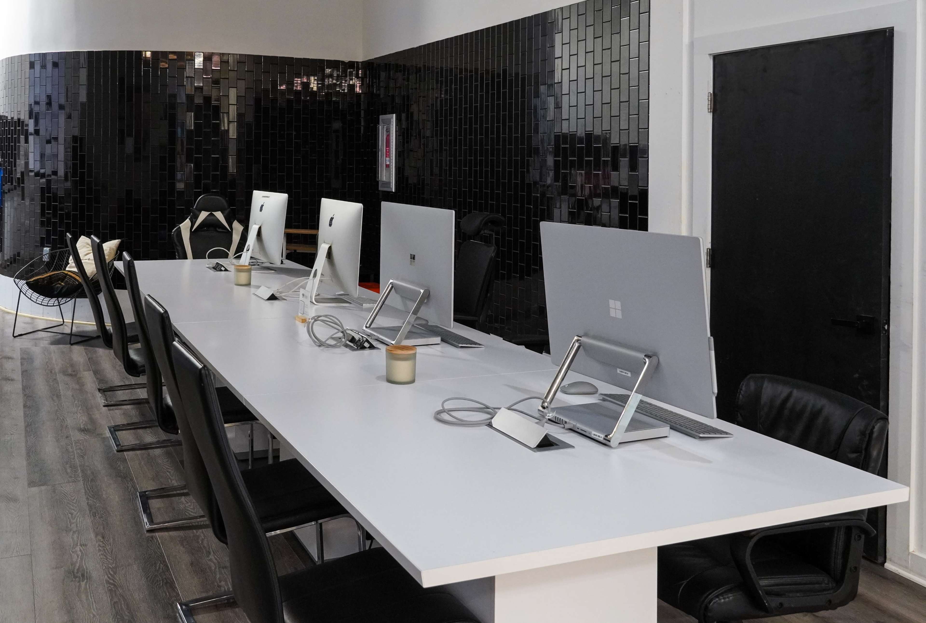 A row of computers is set up on a white table in a modern office space, accompanied by black chairs and a dark wall in the background.