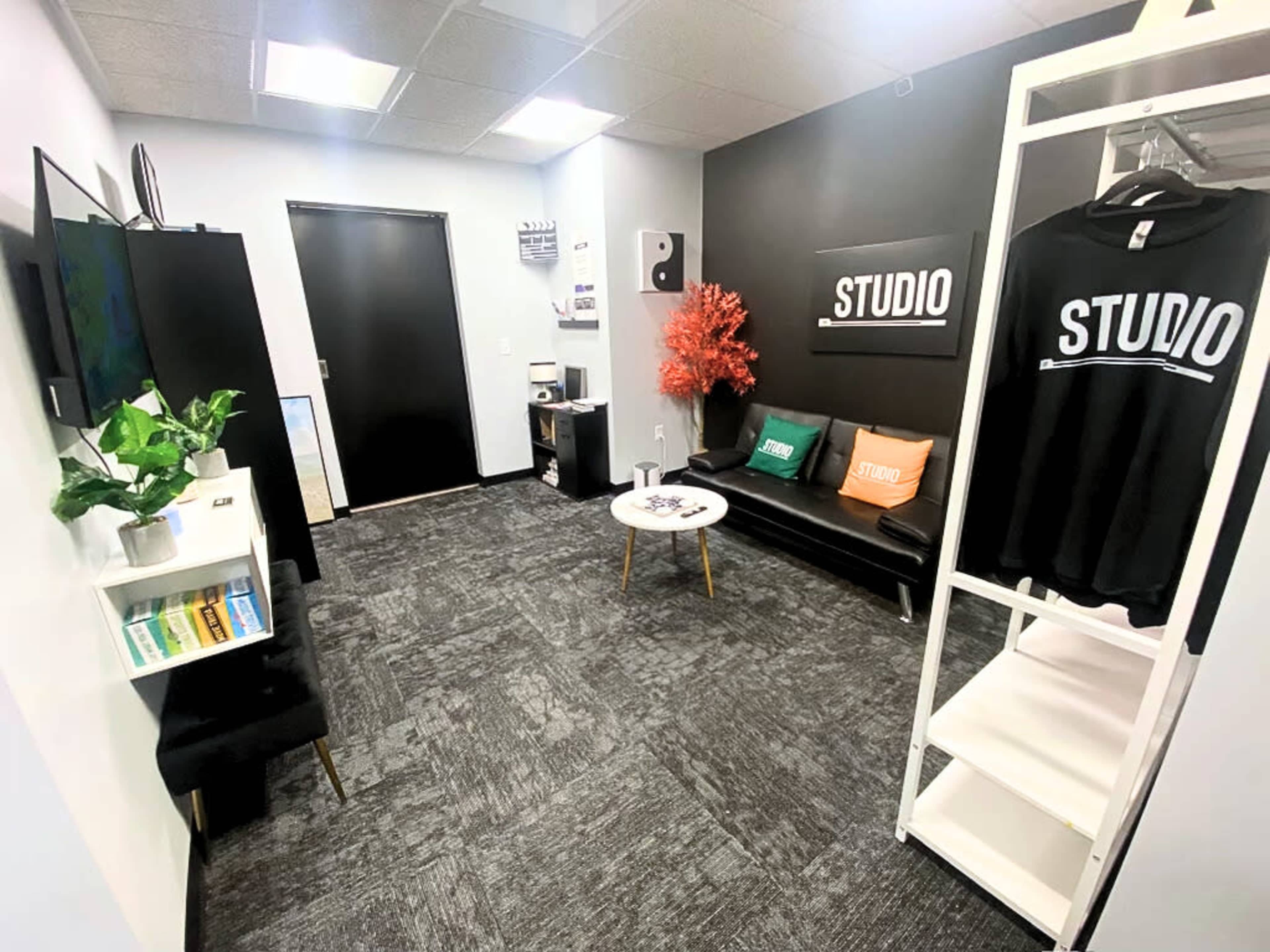 A small studio space with a black accent wall, a seating area with a black couch, a table, and shelving displaying merchandise and plants.