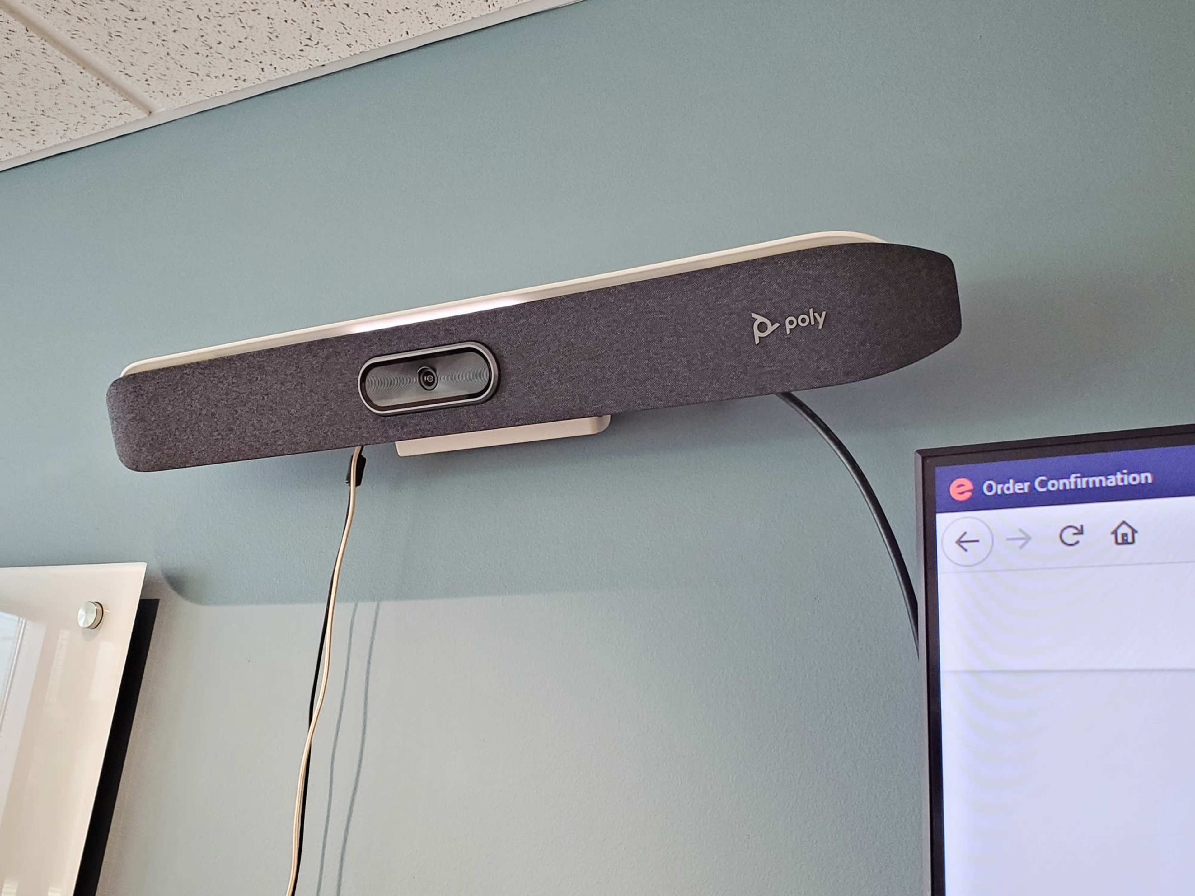 A Poly video conferencing device is mounted on a wall above a computer screen displaying an order confirmation.