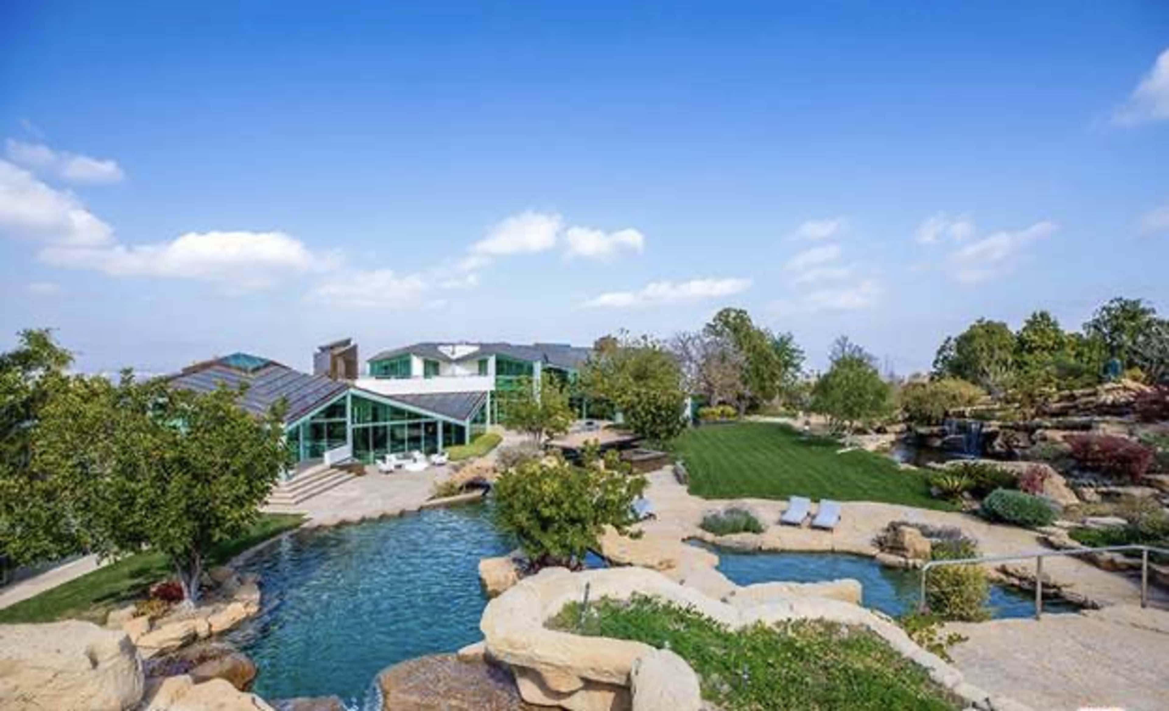 A modern house with large glass windows surrounded by landscaped gardens, a swimming pool, and rocky features under a clear blue sky.
