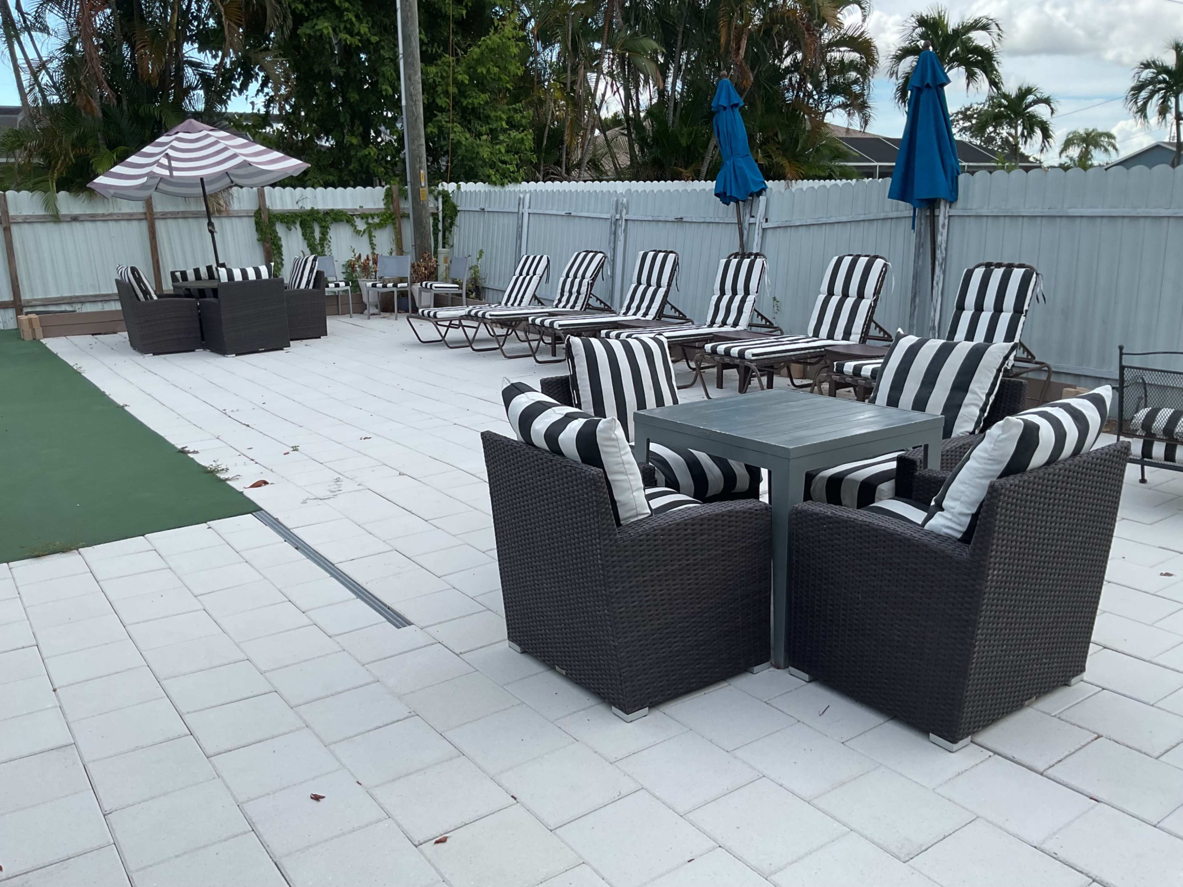 The image shows a poolside area with black and white striped sun loungers, a set of dark wicker chairs and a table, and umbrellas for shade.