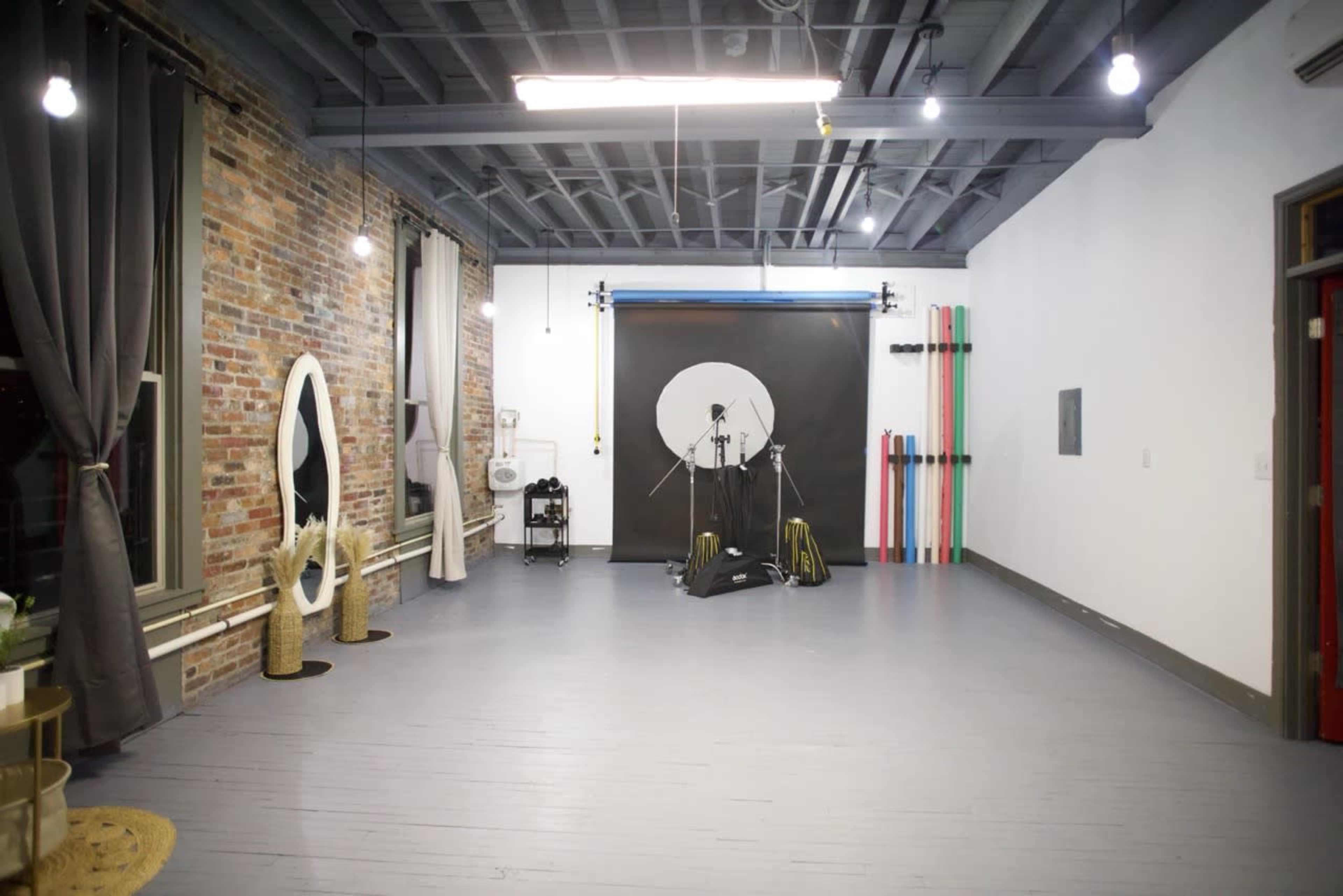 The image shows a spacious studio with exposed brick walls, wooden floors, and various lighting equipment in a minimalistic setup.