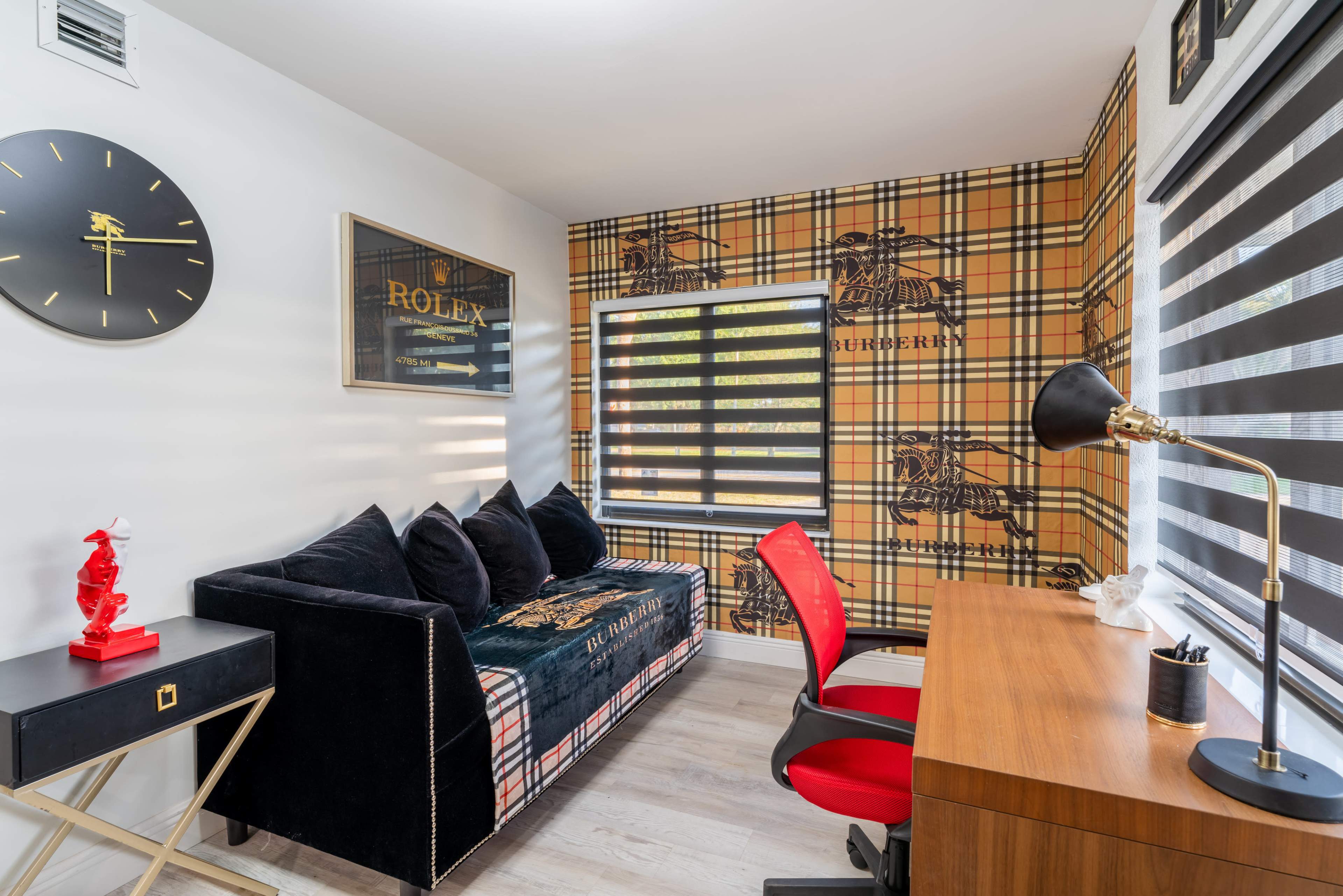 The image shows a stylish office space featuring a plaid wallpaper, a black couch, a wooden desk with a lamp, and a large window with striped blinds.