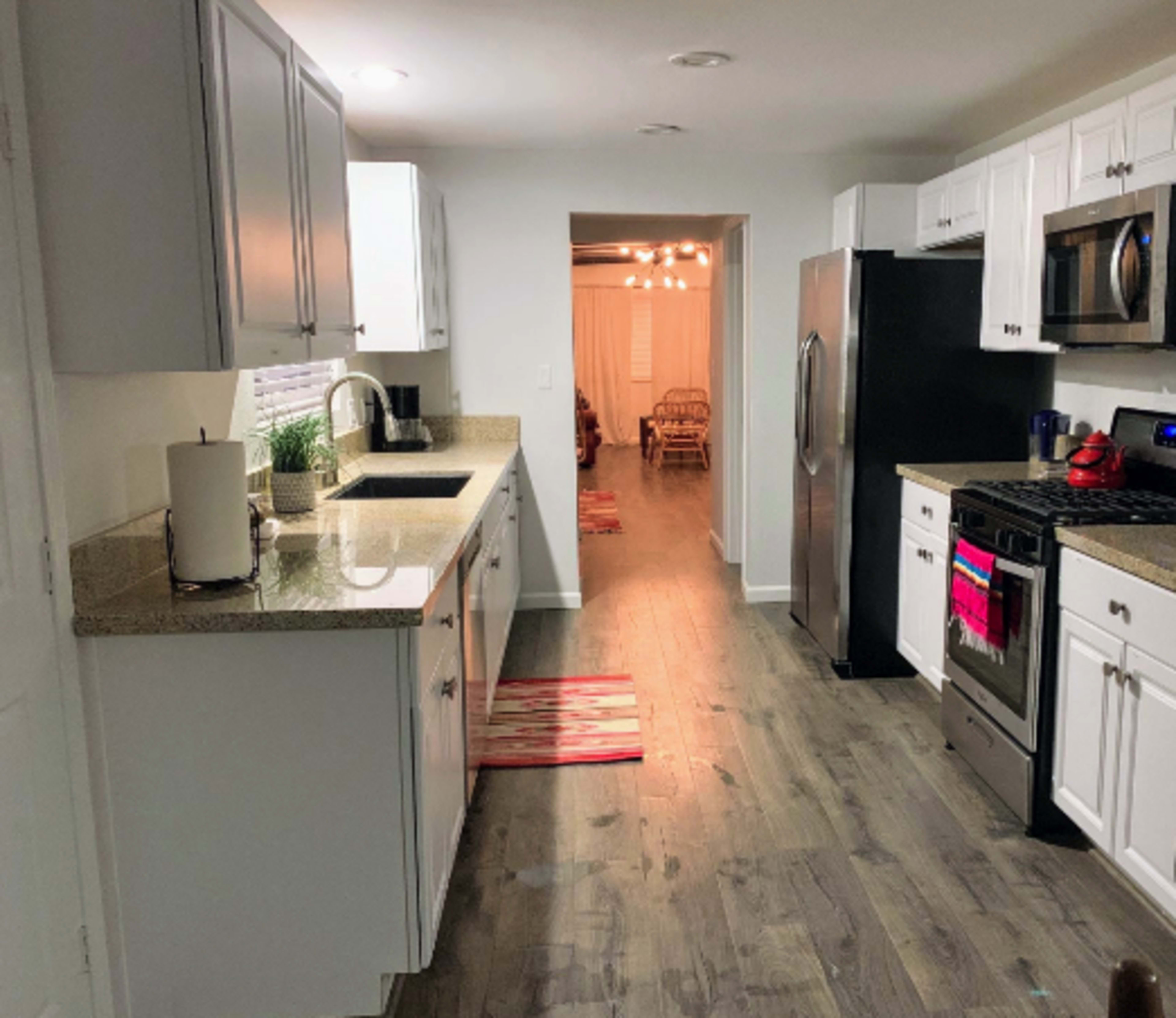 The image shows a modern kitchen with white cabinets, a granite countertop, stainless steel appliances, and a view into a well-lit dining area.
