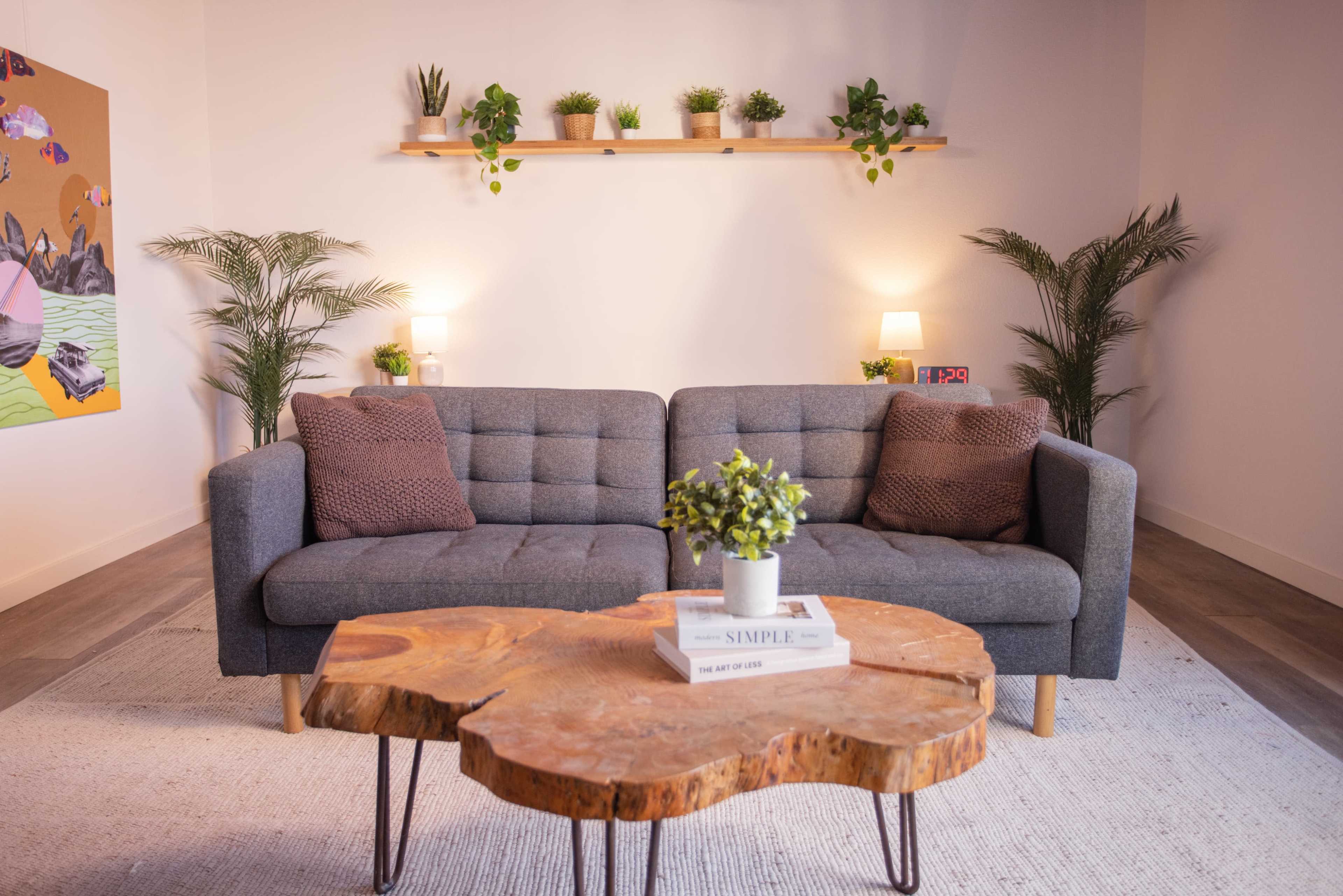 The image shows a modern living room with a gray sofa, a wooden coffee table, and decorative plants on shelves and the floor.