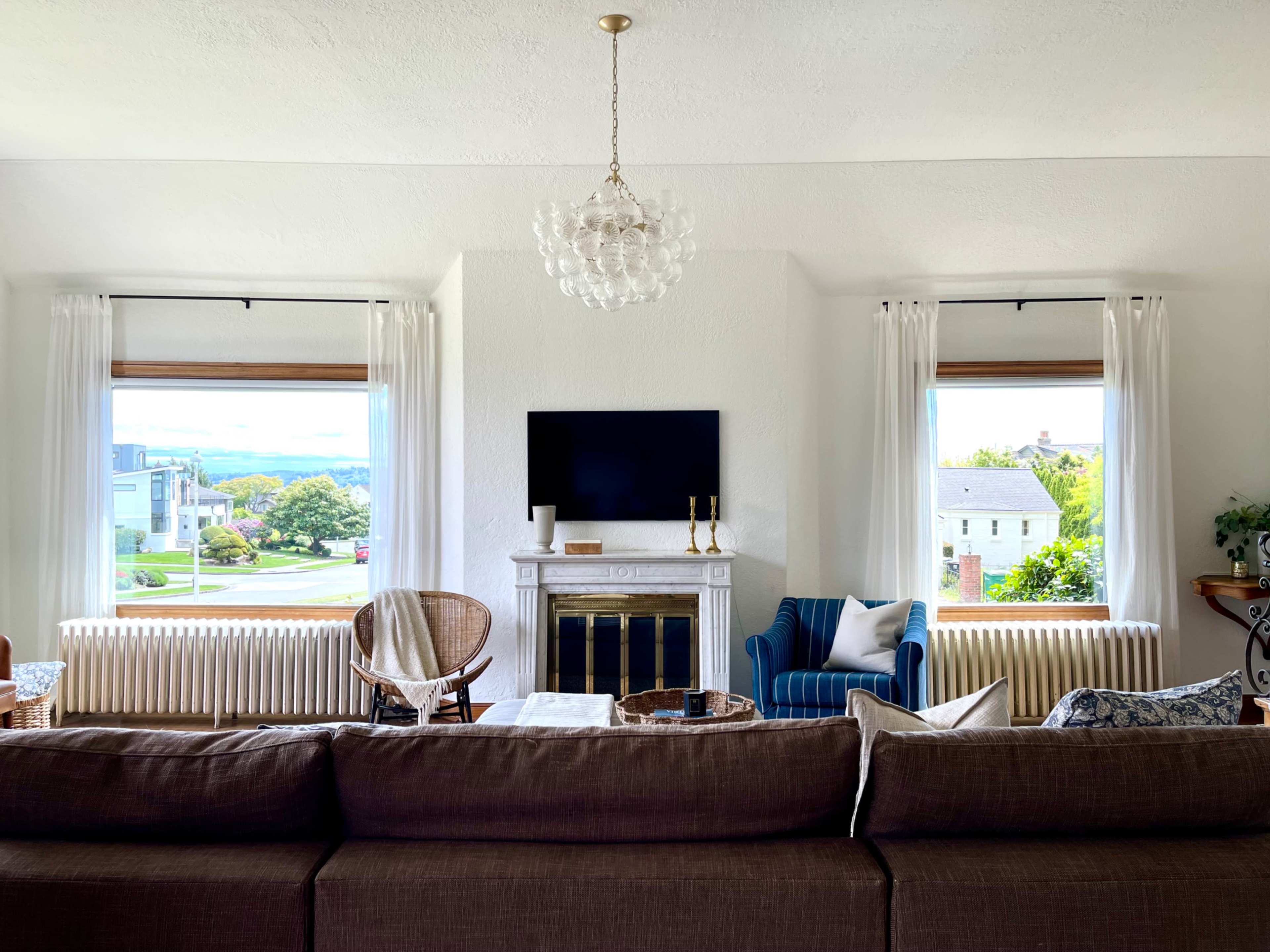A living room features a fireplace with a television above it, two windows with curtains, a sofa, a blue armchair, and a decorative table in front.