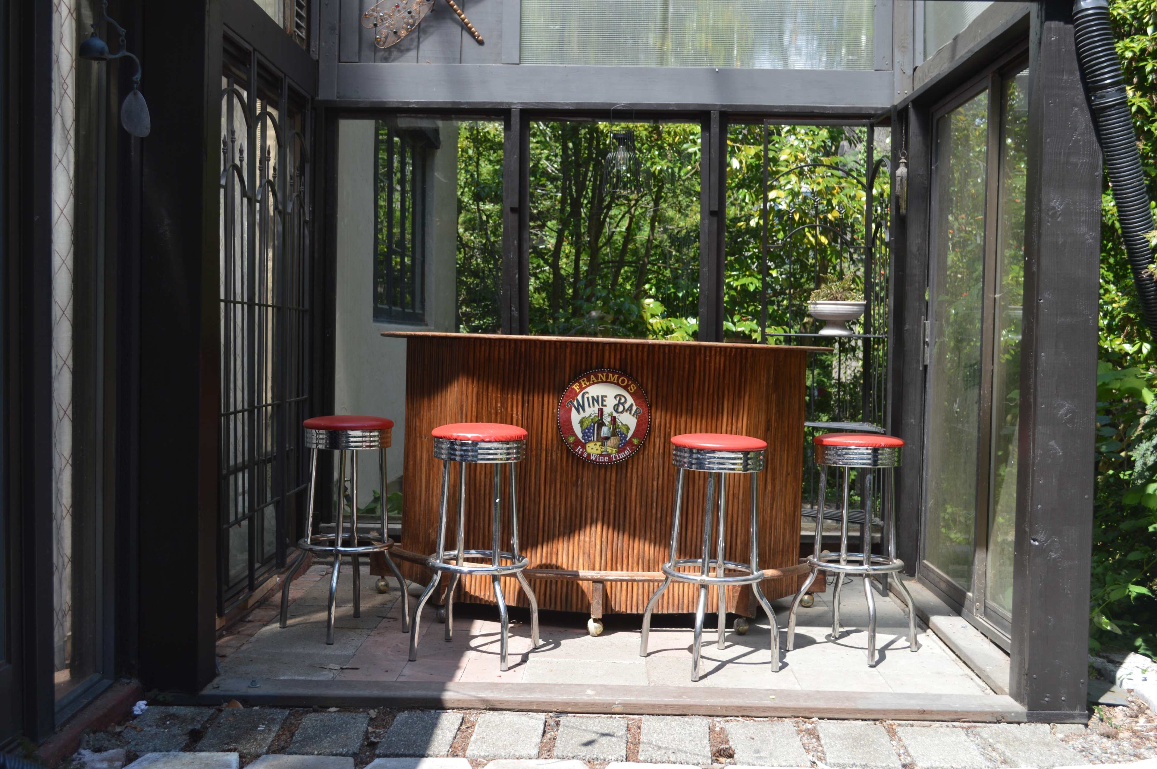A small outdoor bar with a wooden counter and four metal stools, situated in a glass-enclosed area surrounded by greenery.