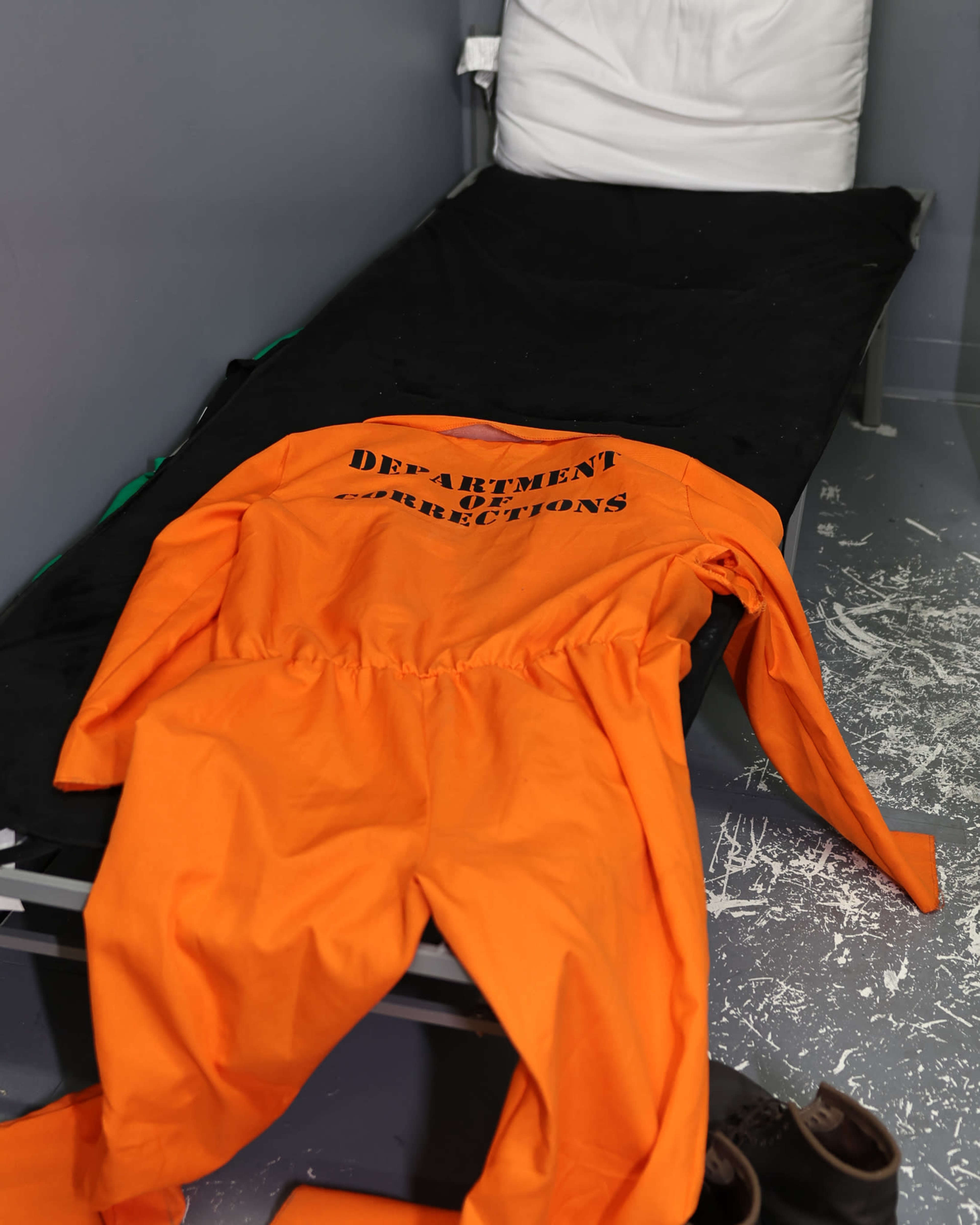An orange jumpsuit labeled "Department of Corrections" is laid out on a cot in a gray room.