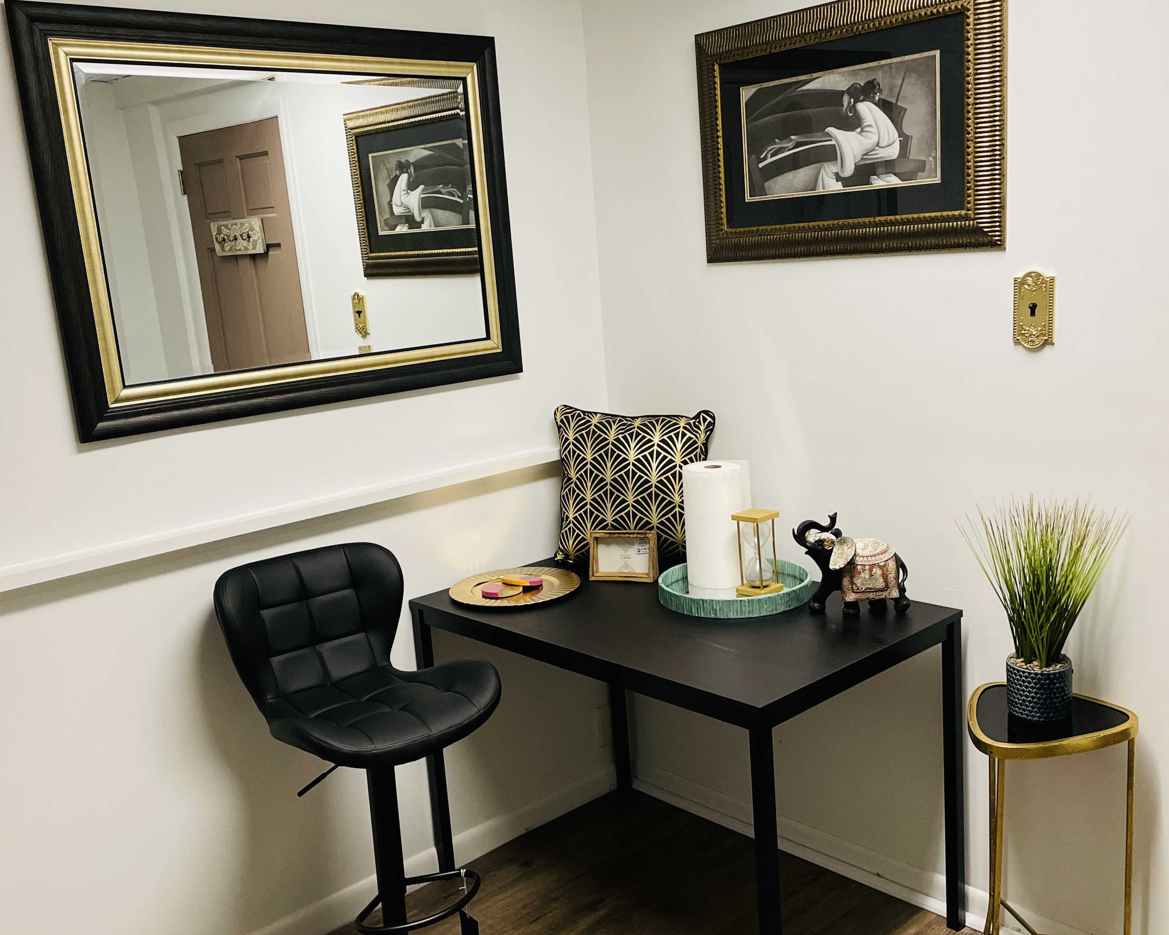 The image shows a small corner of a room featuring a black desk with a stool, decorative items on the desk, and framed artwork on the walls.
