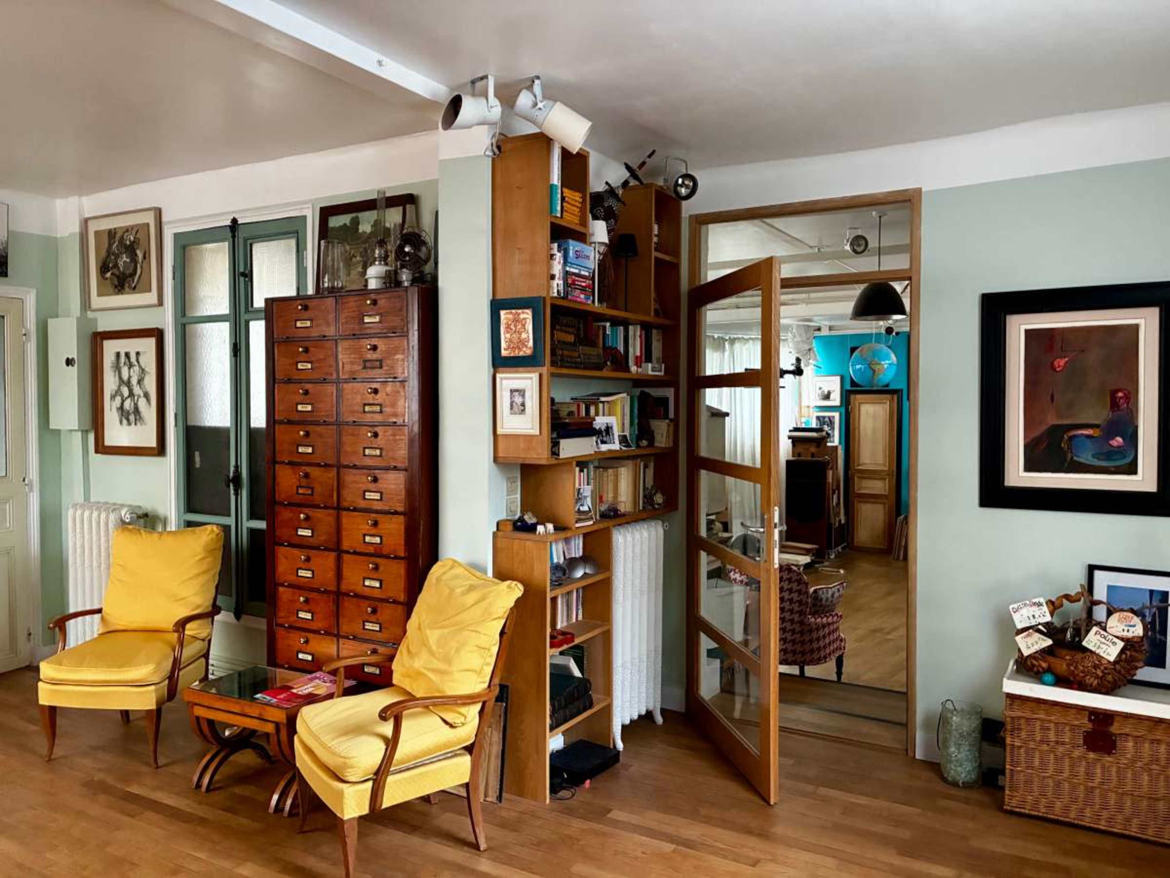 The image shows a stylish room with yellow armchairs, a wooden bookshelf, and an open door leading to another space adorned with various artworks and furniture.
