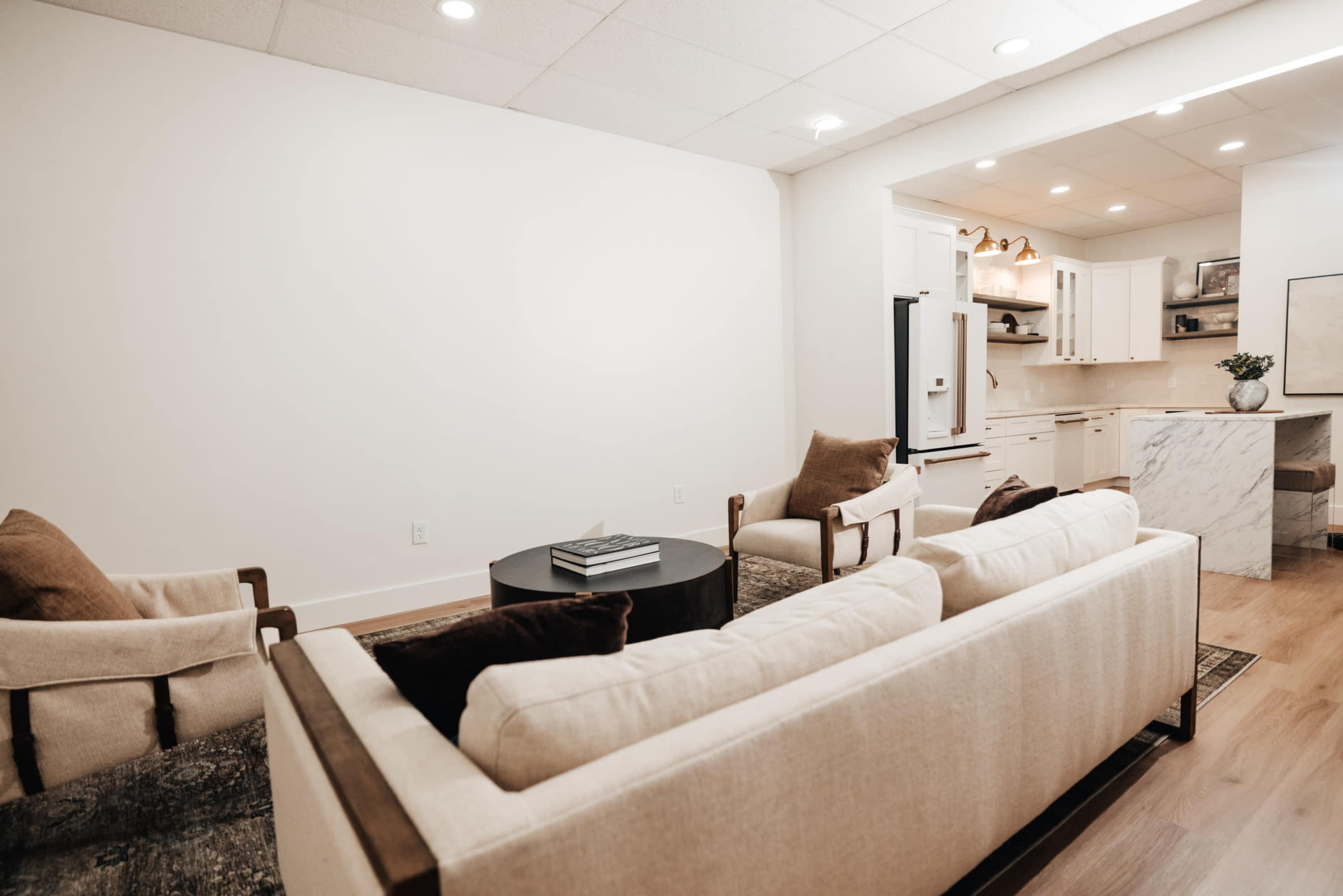 A modern living room with a beige sofa and armchairs arranged around a dark coffee table, adjacent to a light-colored kitchen area.