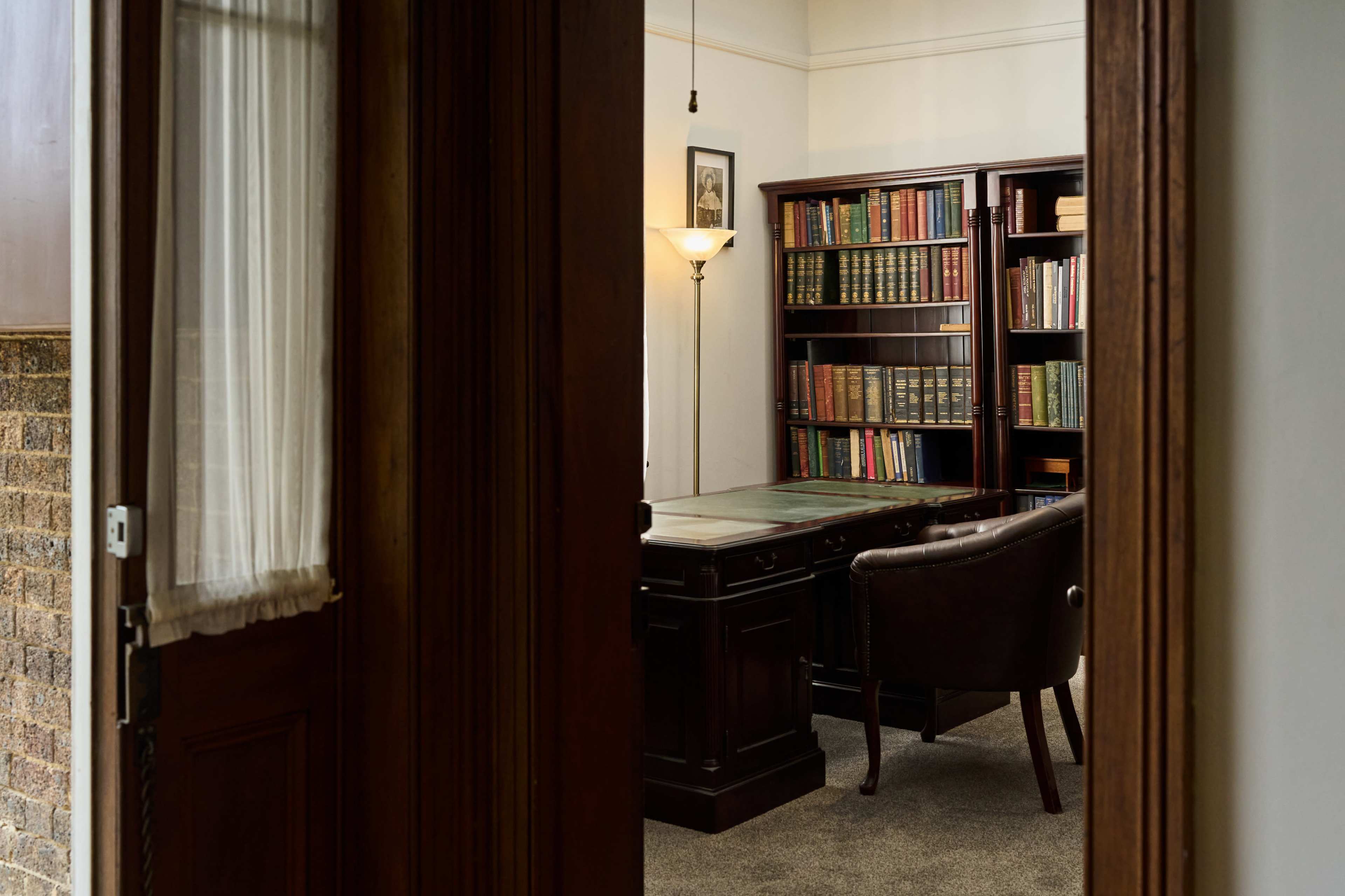 The image shows a well-appointed office with a wooden desk, a chair, and a bookshelf filled with books, as seen through an open door.