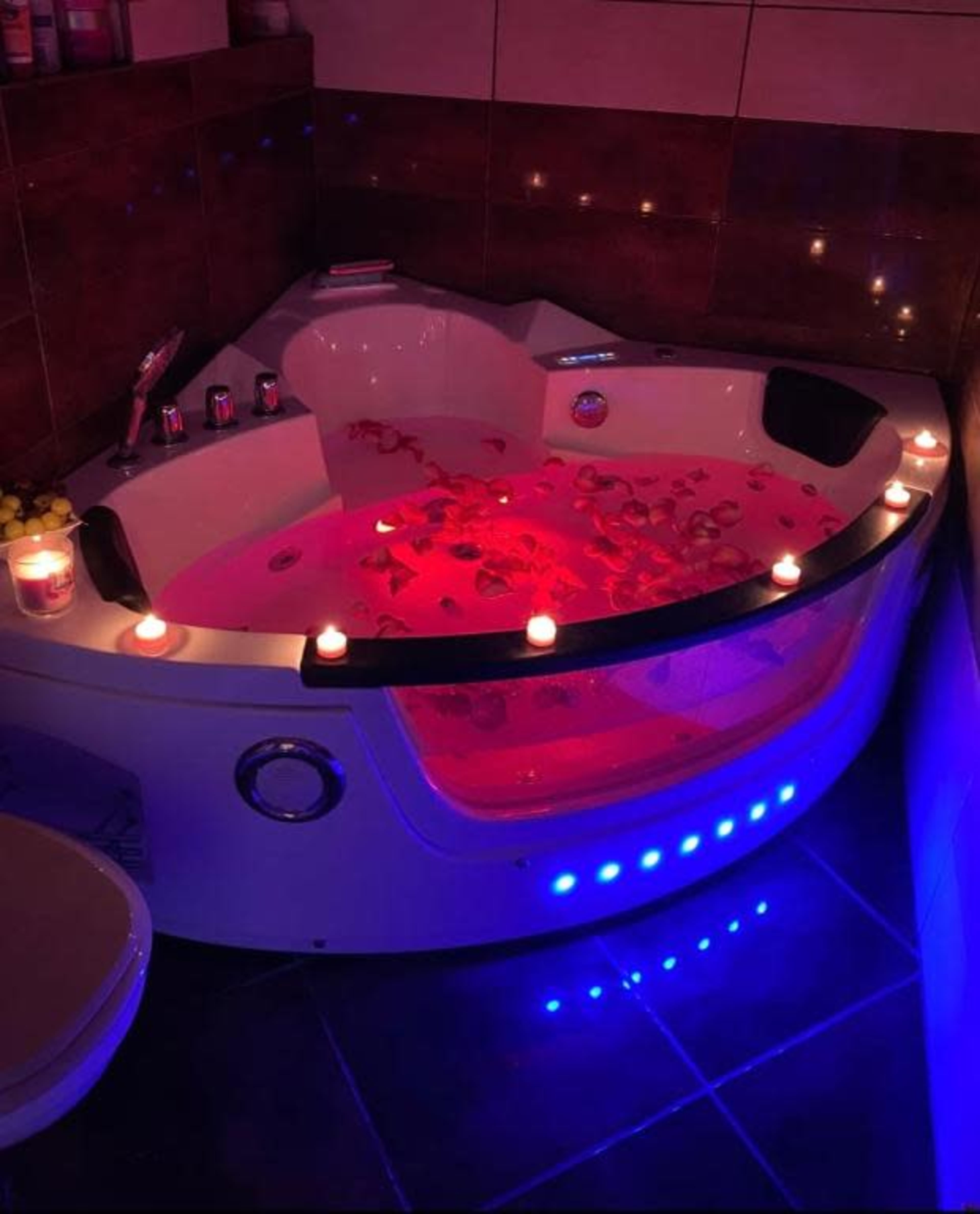 The image shows a Jacuzzi filled with water and rose petals, illuminated by pink and blue lights, surrounded by candles.