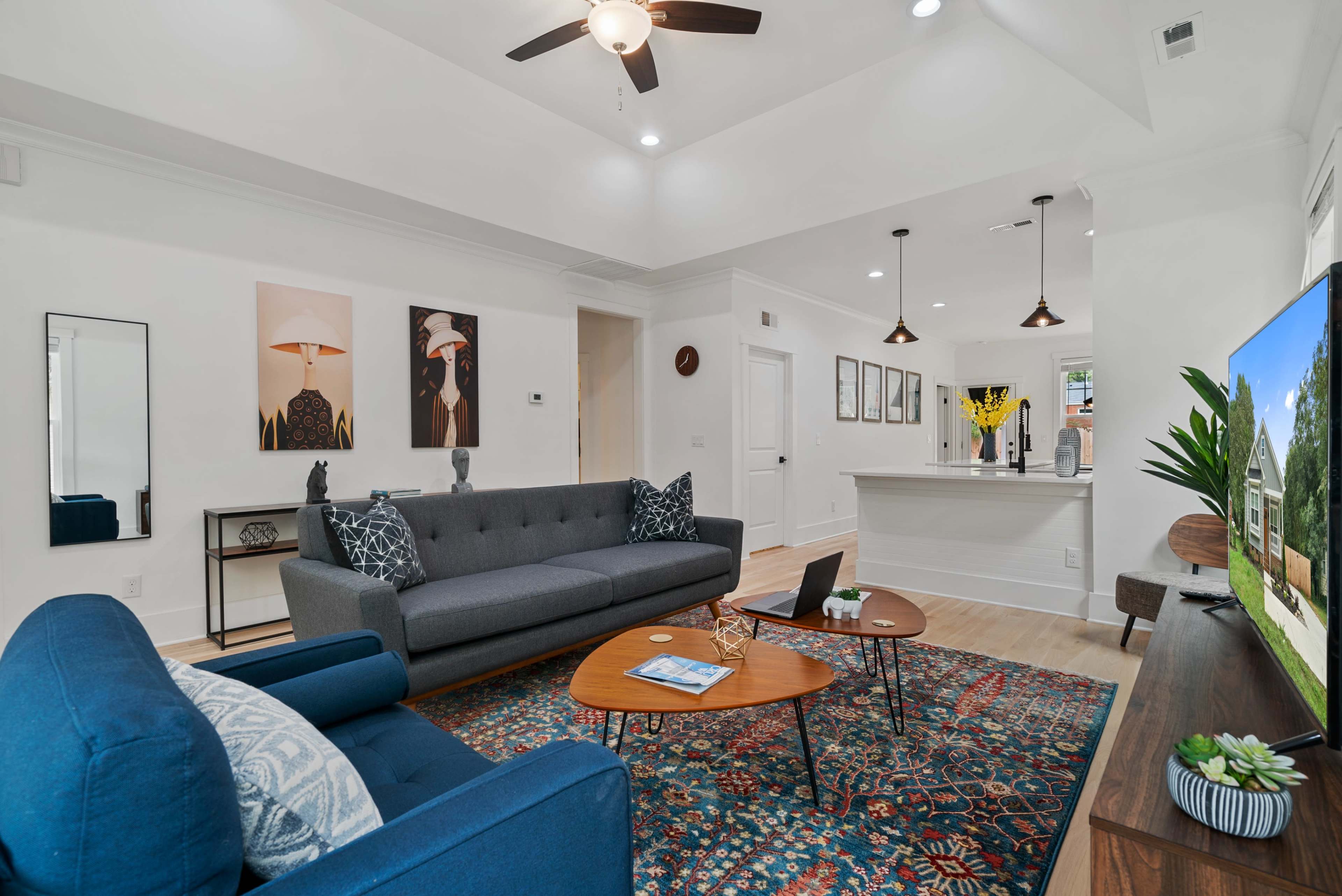 The living room features a gray sofa and a blue armchair, arranged around a coffee table on a decorative rug, with artwork on the walls and a ceiling fan above.