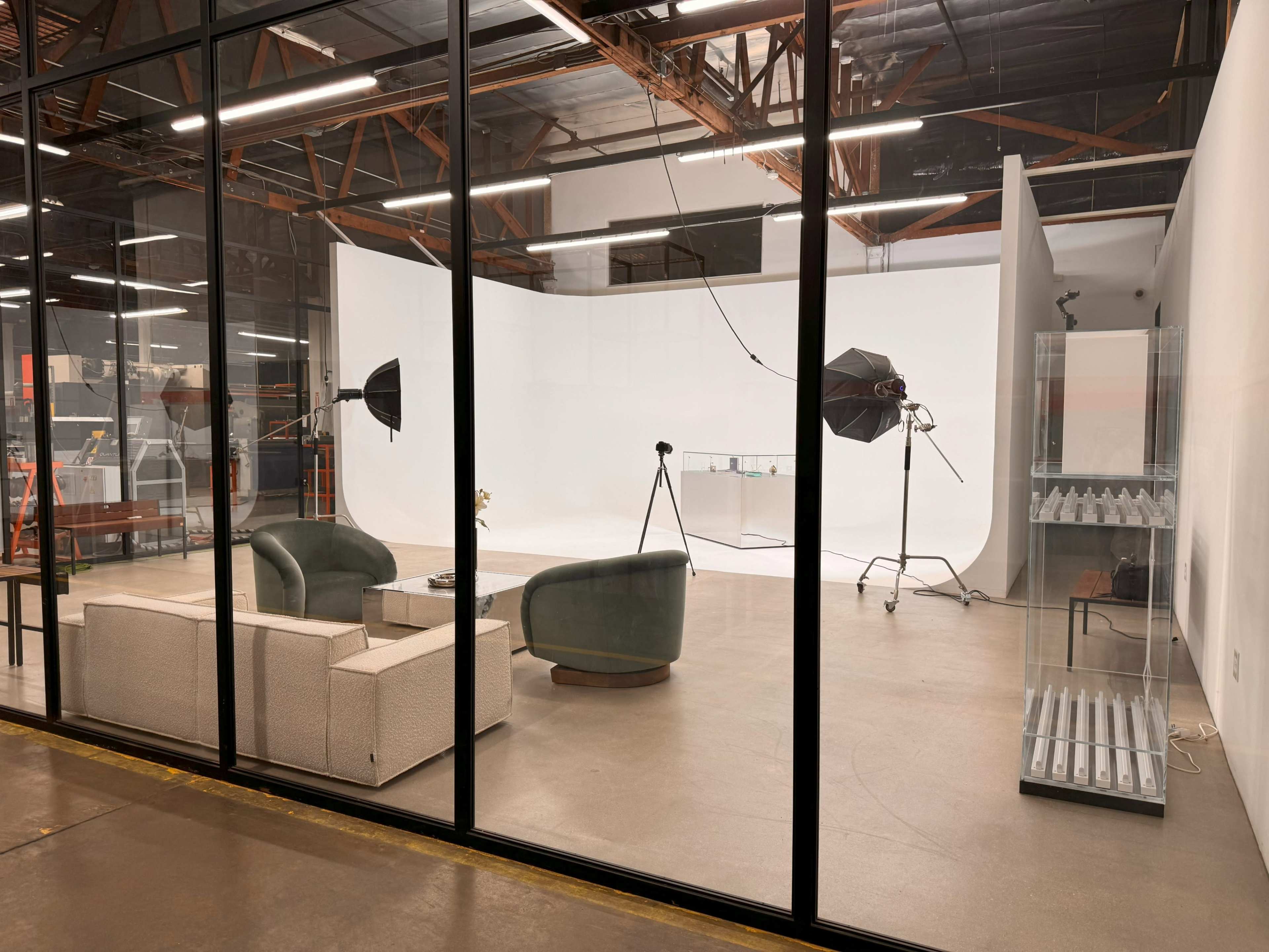 The image shows a photography studio featuring a minimalist setup with a white backdrop, studio lighting equipment, and a seating area with two chairs and a coffee table.