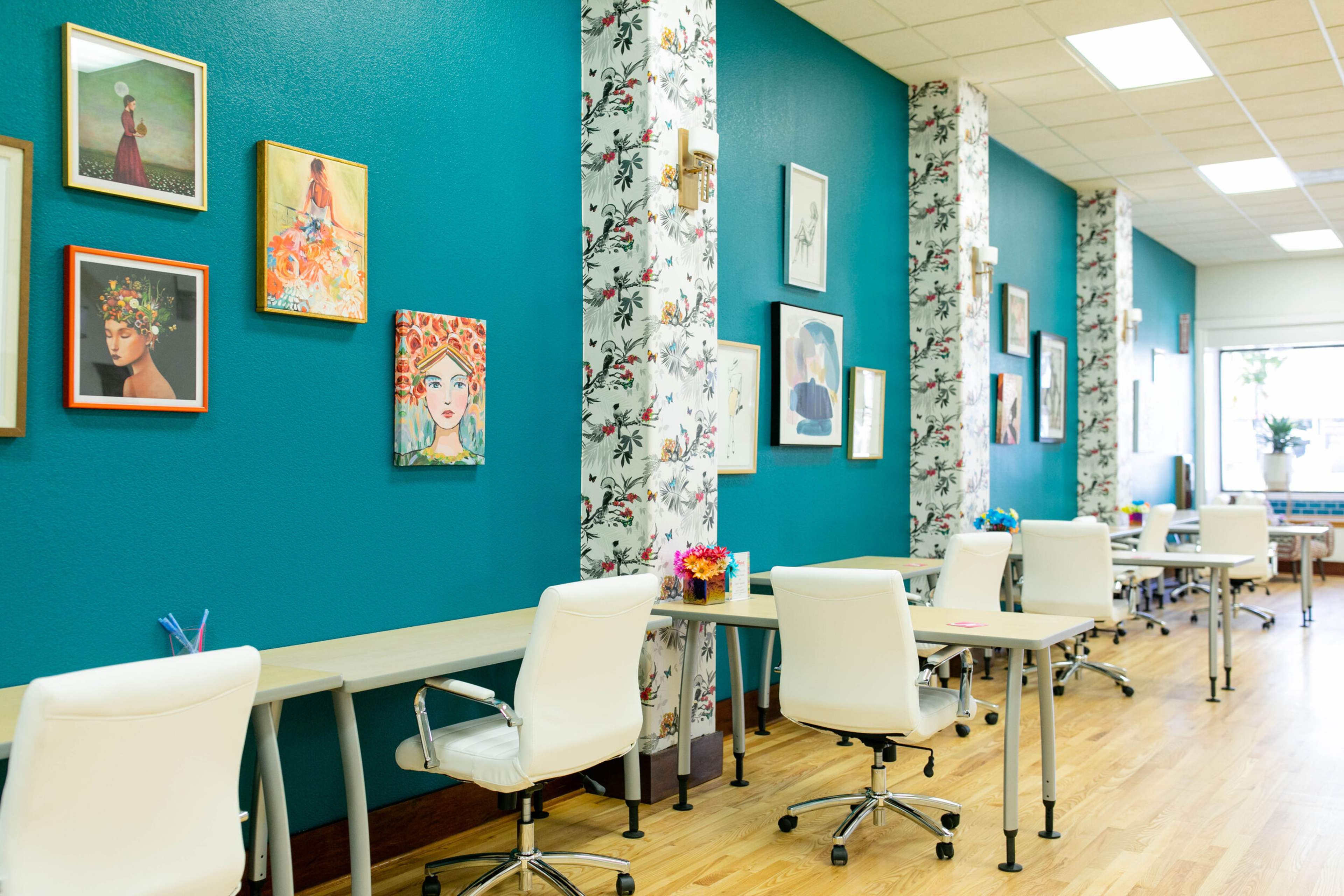 A brightly colored workspace with teal walls, floral wallpaper, and several art pieces hanging on the walls, alongside rows of white chairs and desks.