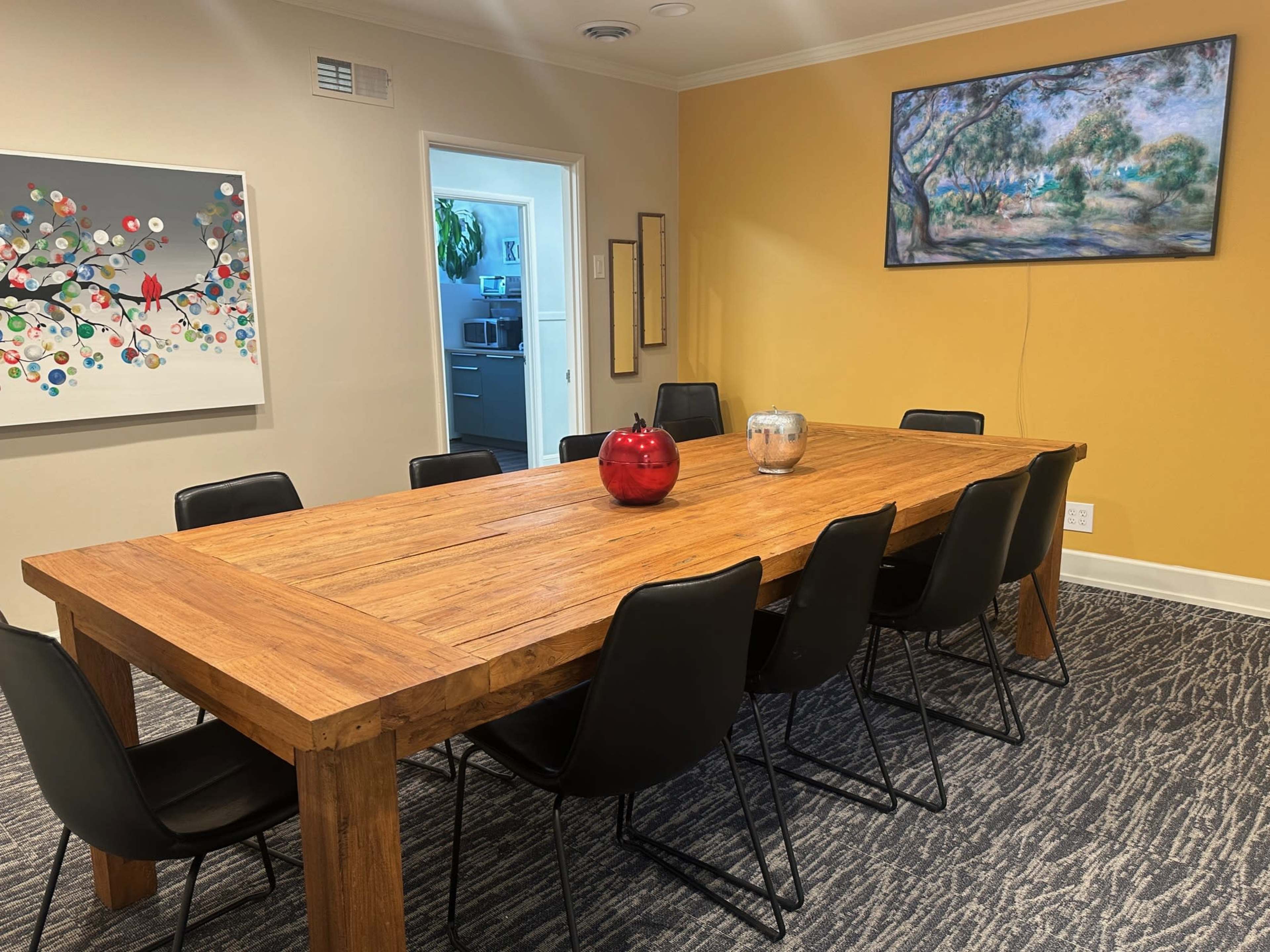 A large wooden dining table surrounded by black chairs is set in a room with an orange wall and a painting hanging above it, while an open doorway leads to a kitchen area.
