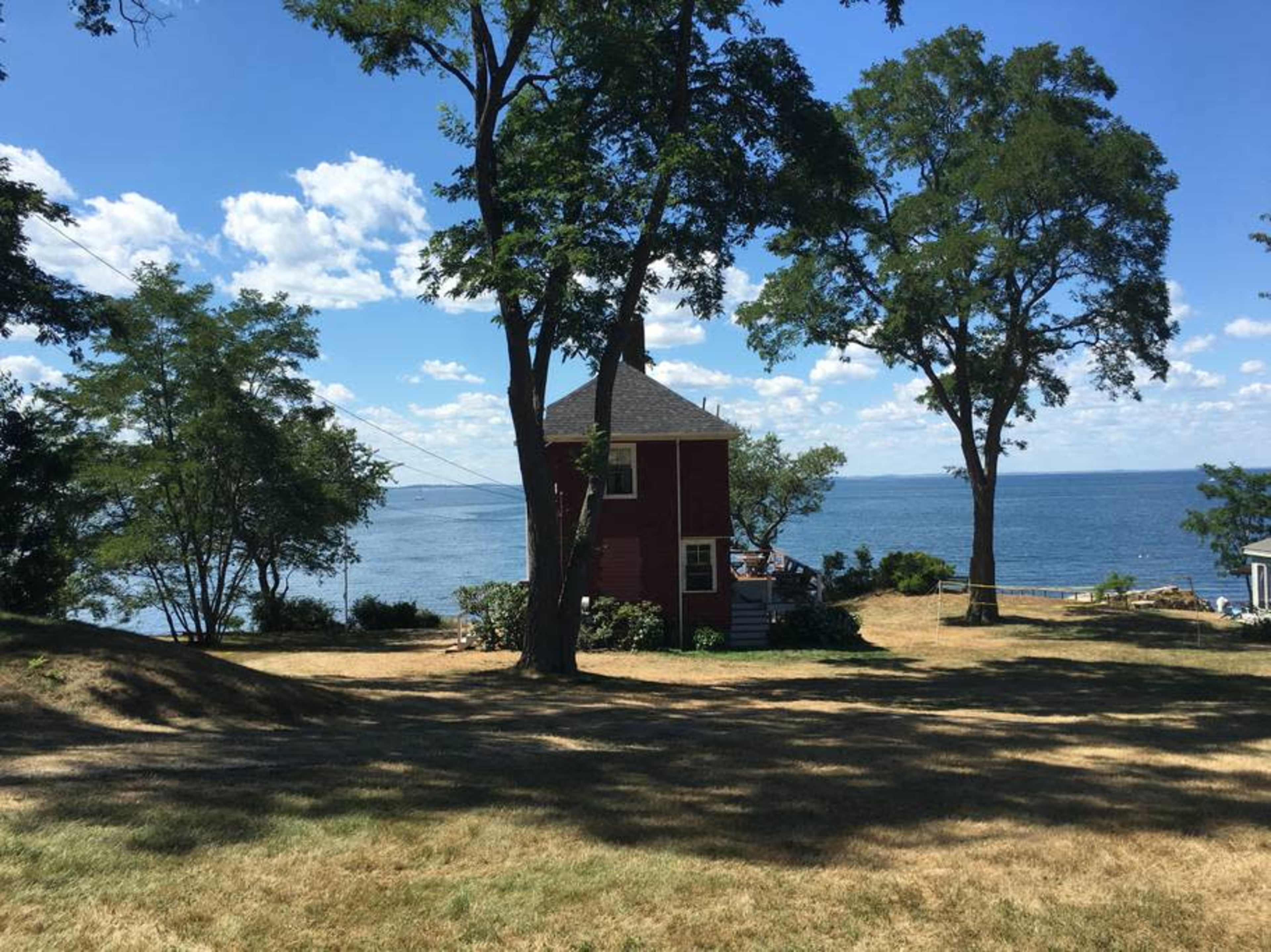 A red house stands beside a calm body of water, surrounded by trees and grassy land under a blue sky.