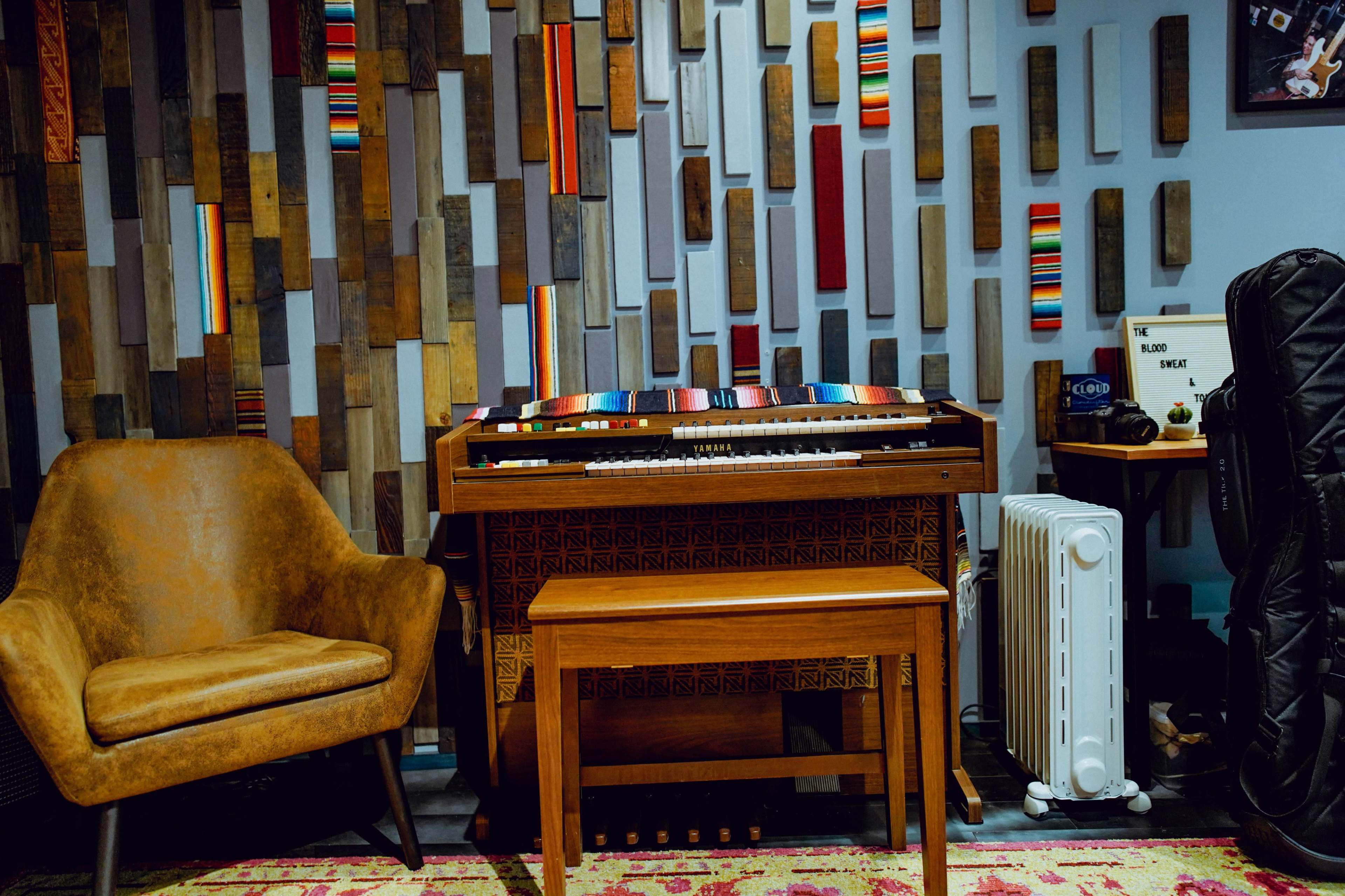 The image shows a cozy music room featuring a wooden organ with a matching bench, an armchair, and a patterned rug, all set against a colorful wooden accent wall.