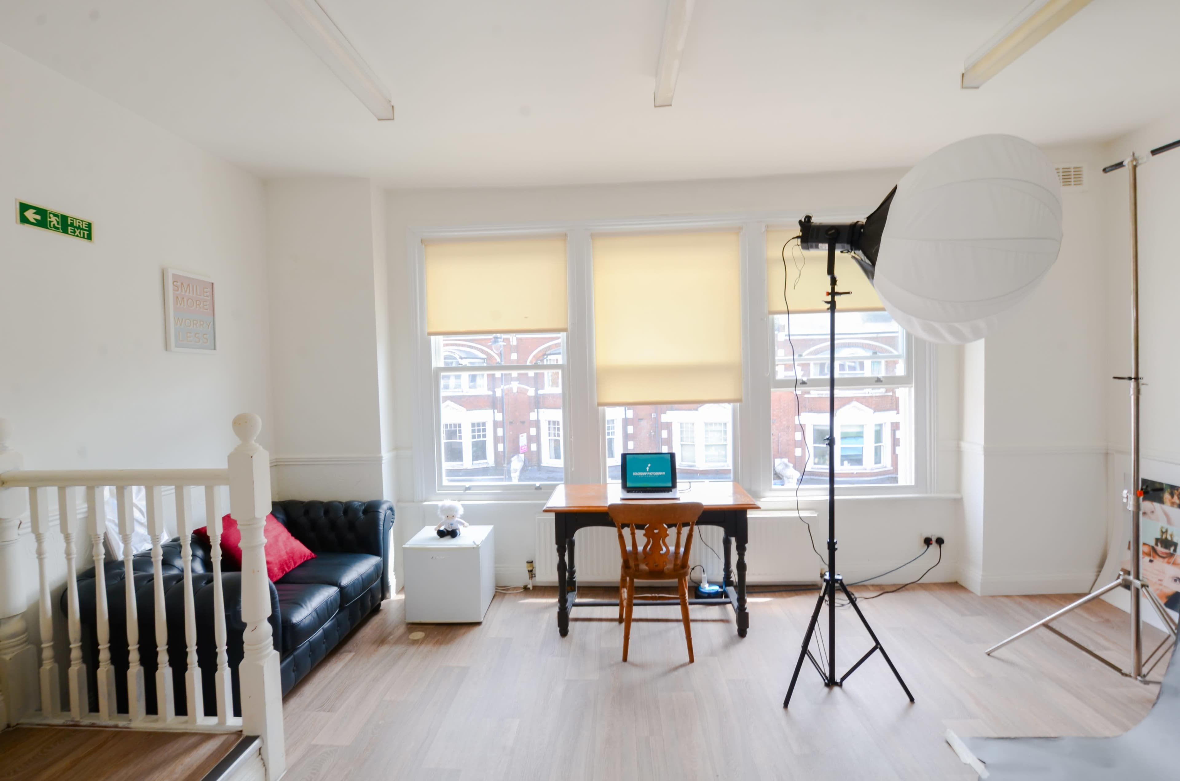 The image shows a bright room with large windows, featuring a black sofa, a wooden table with a laptop, and a photography light setup.