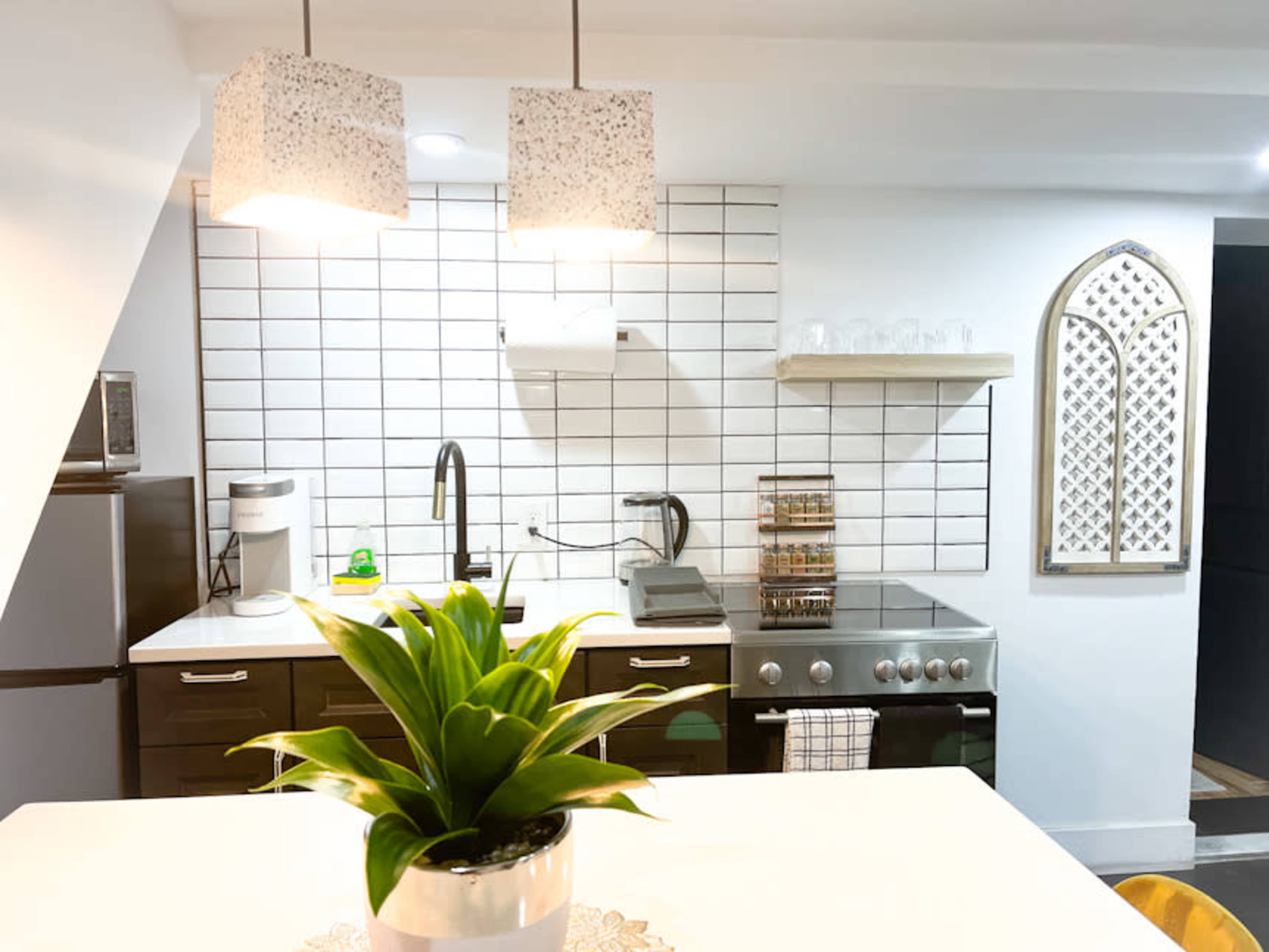 A modern kitchen with sleek dark cabinets, a white tiled backsplash, a stainless steel stove, and pendant lights hanging above a dining table with a decorative plant.