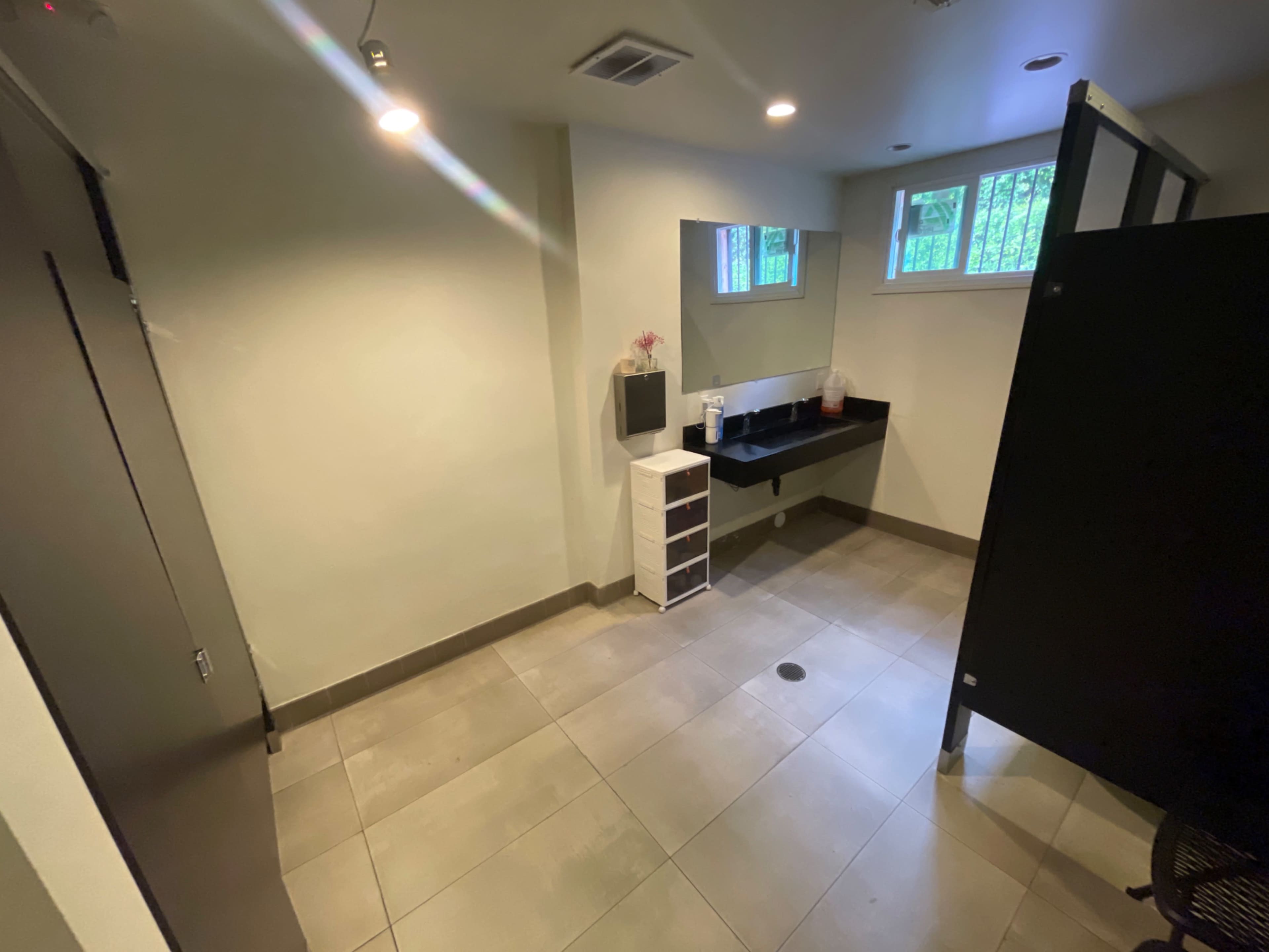 The image shows a clean restroom interior with a black countertop, a mirror, a small cabinet, and a privacy stall, all arranged on tiled flooring.