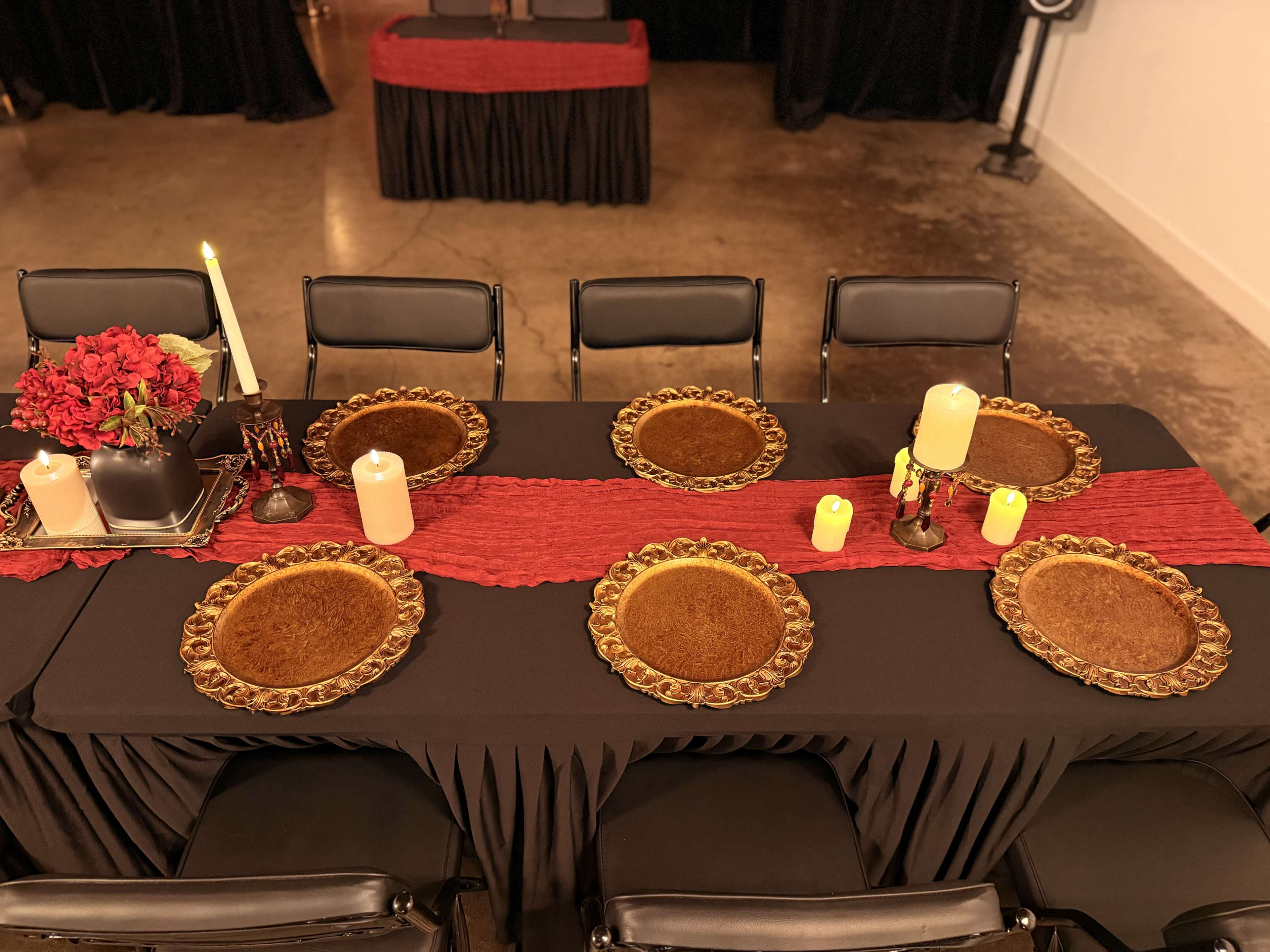 A long dining table is set with round brown plates, candles, and a centerpiece of red flowers, surrounded by black chairs.