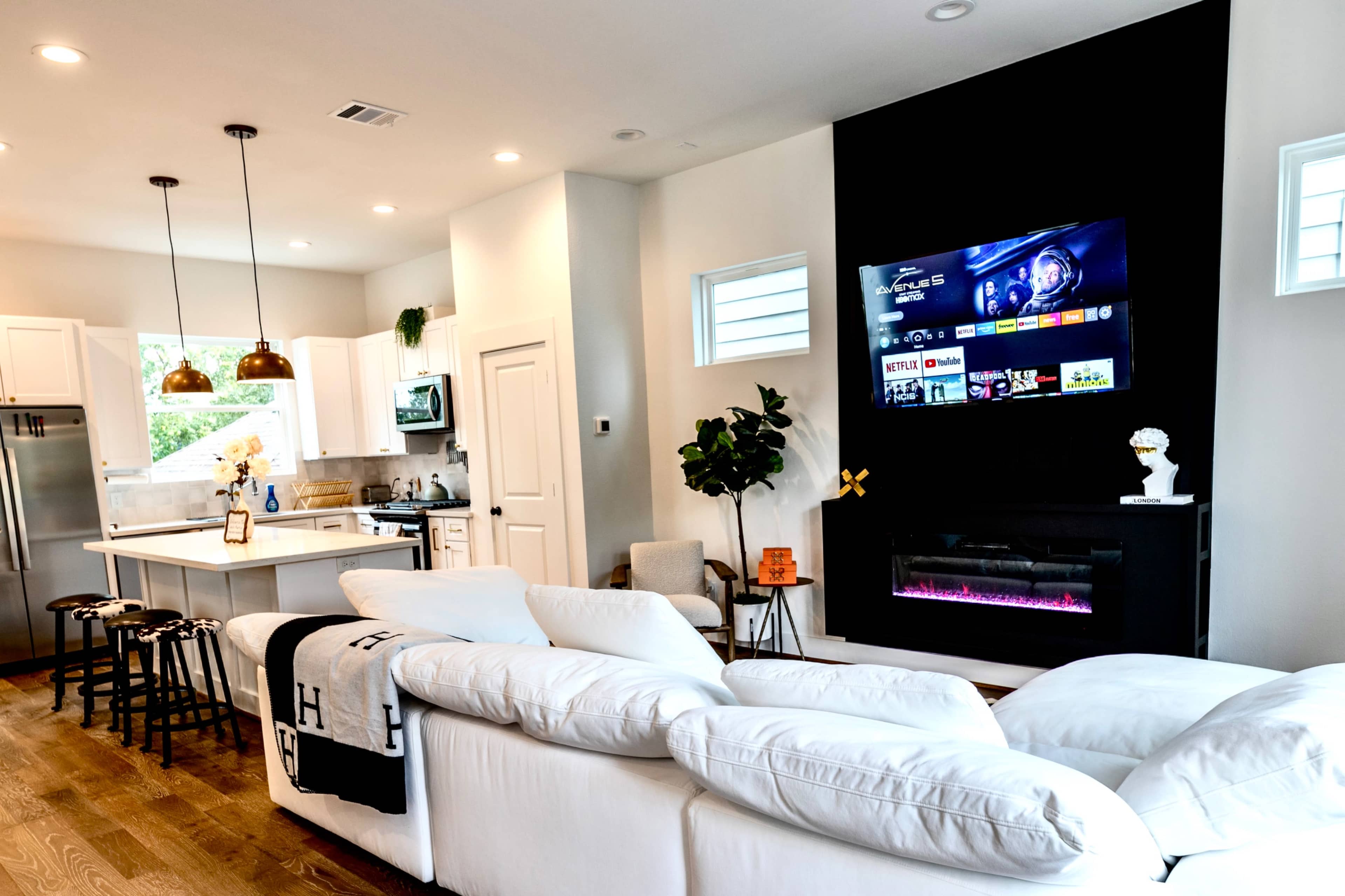 A modern living room features a white sectional sofa facing a wall-mounted TV above an electric fireplace, with a kitchen and dining area visible in the background.