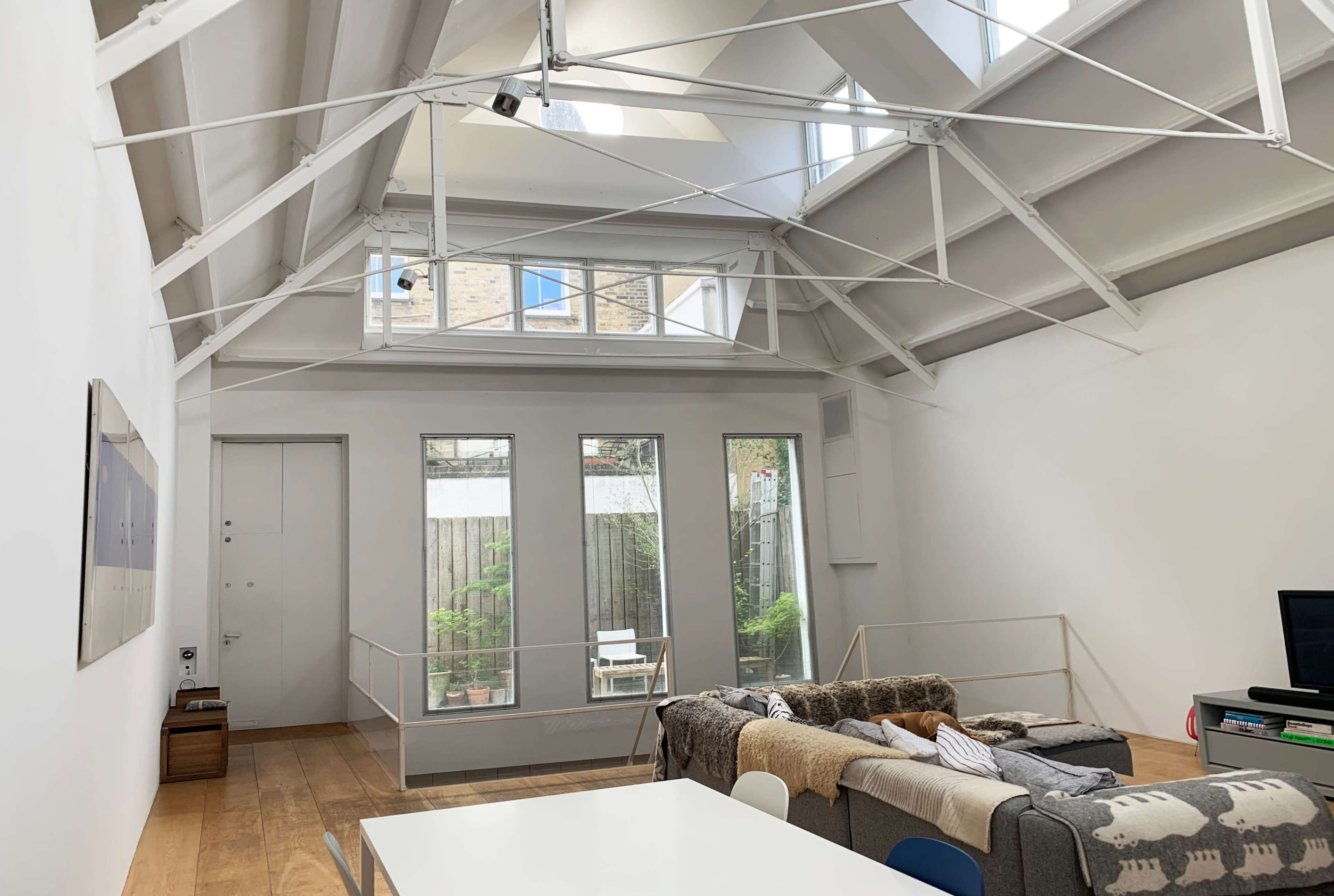 The image depicts a modern, open-concept interior space with a high ceiling, large windows, and a living area featuring a sofa and a dining table.