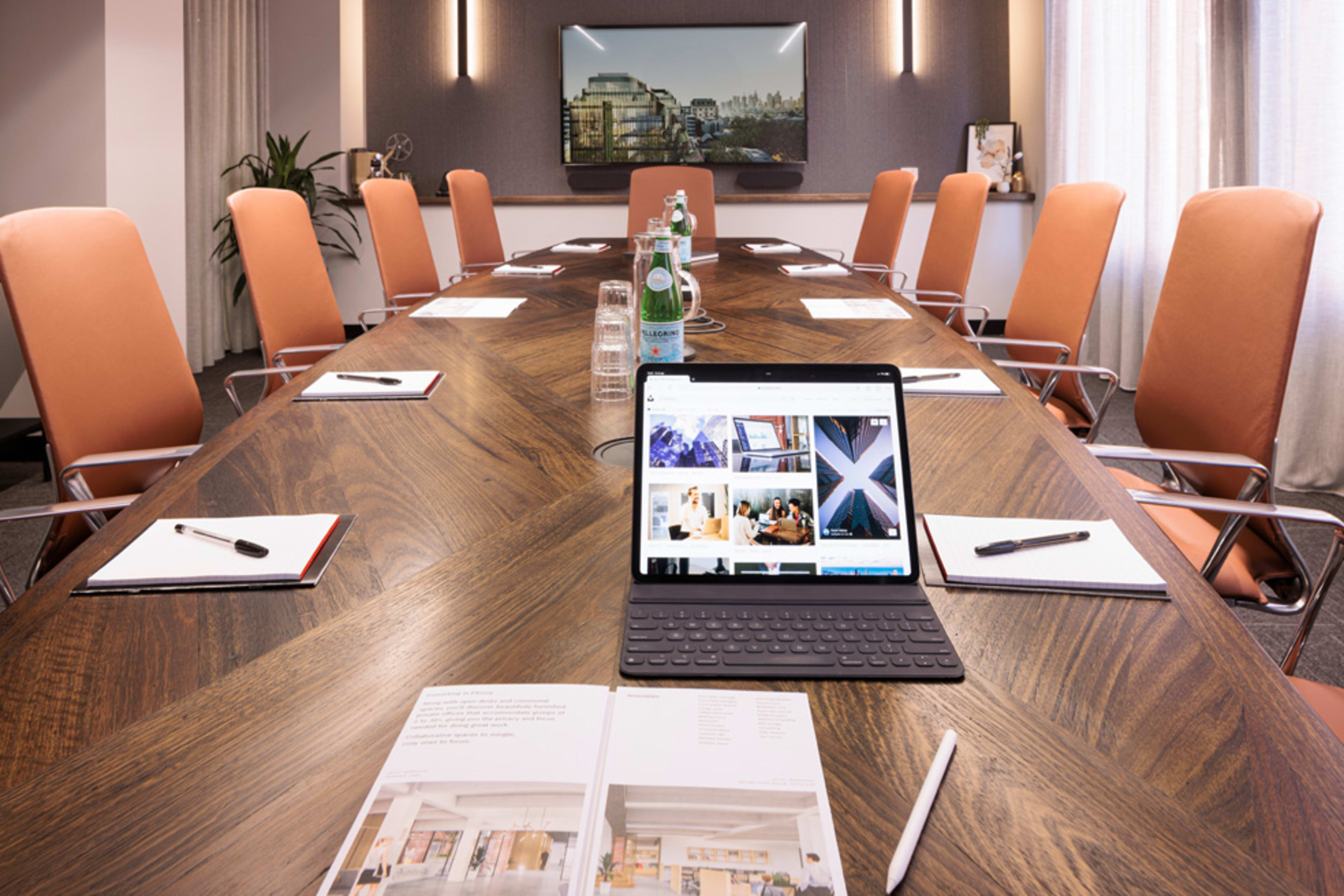 A modern conference room features an elongated wooden table, equipped with stationery and an iPad displaying images, surrounded by orange chairs.
