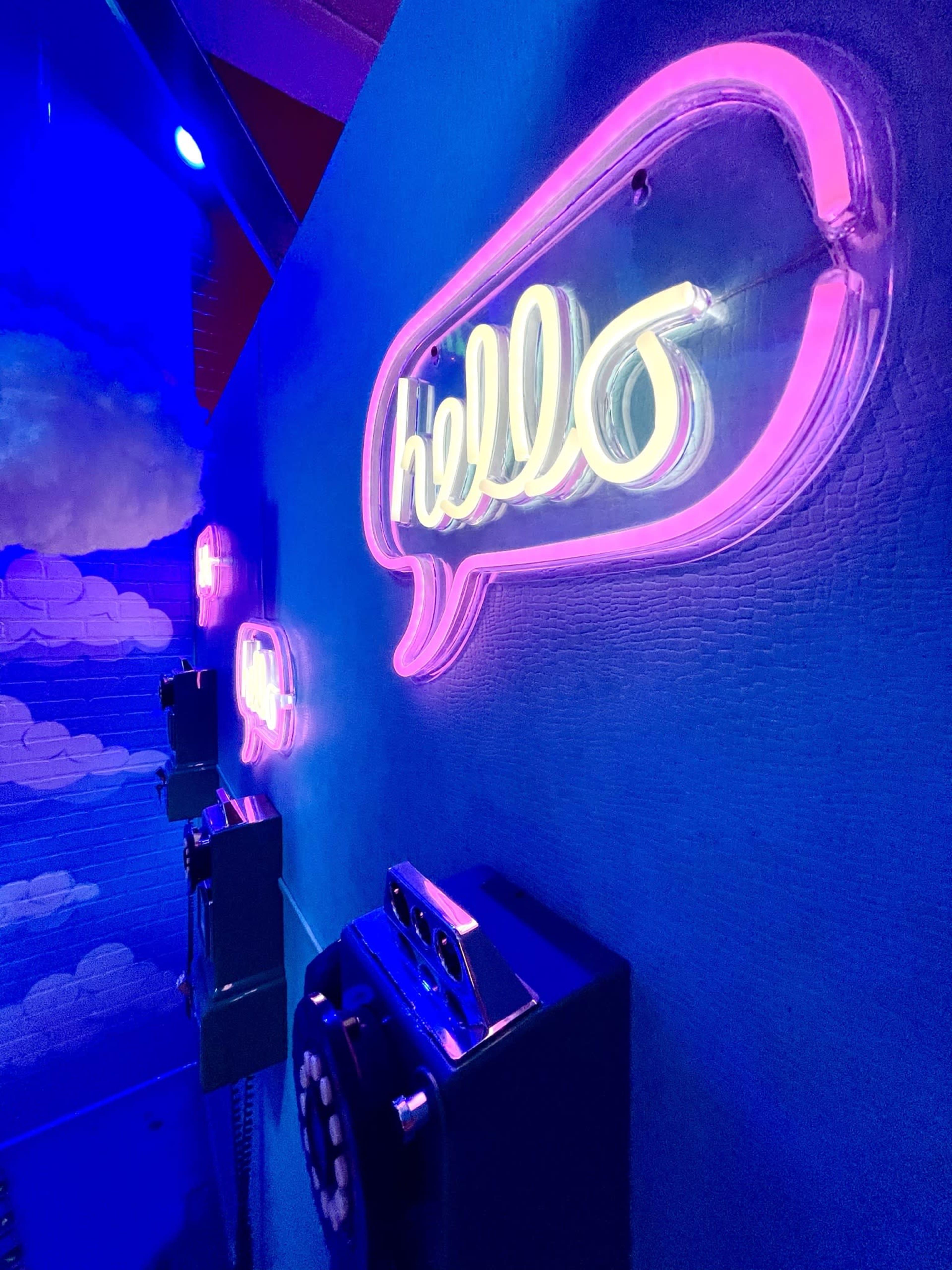 A neon sign in the shape of a speech bubble saying "hello," illuminated on a wall beside vintage telephones.