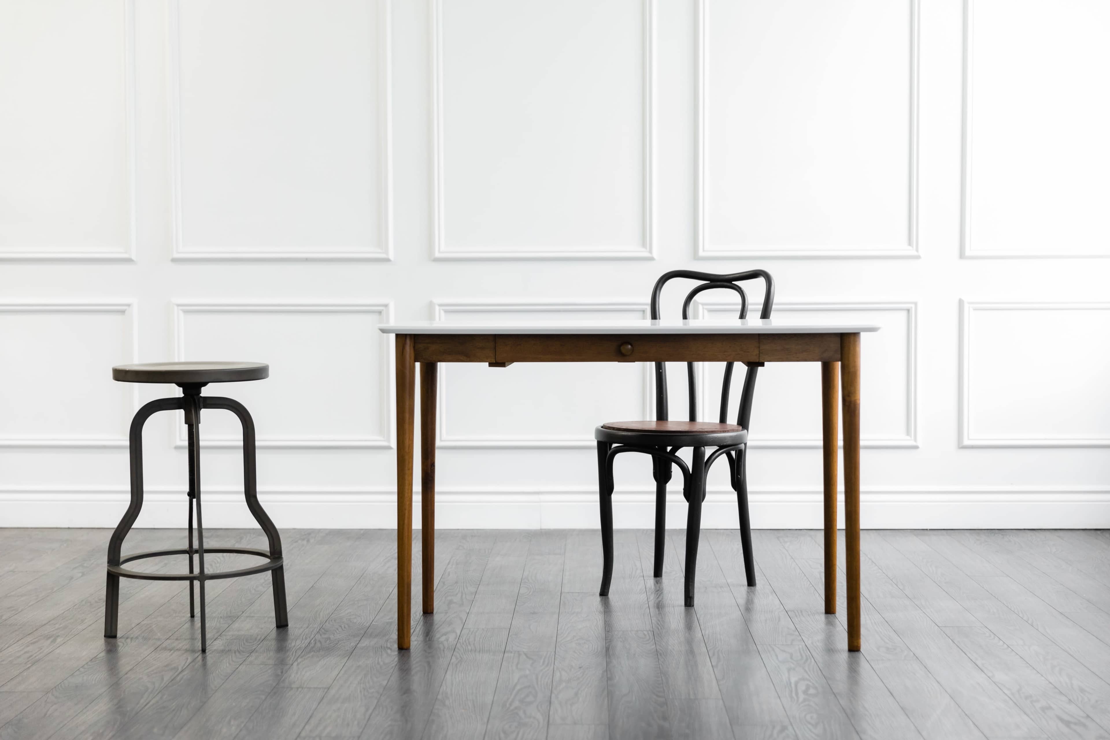 A wooden table and a chair are positioned in a minimalist room with white paneled walls and hardwood flooring.