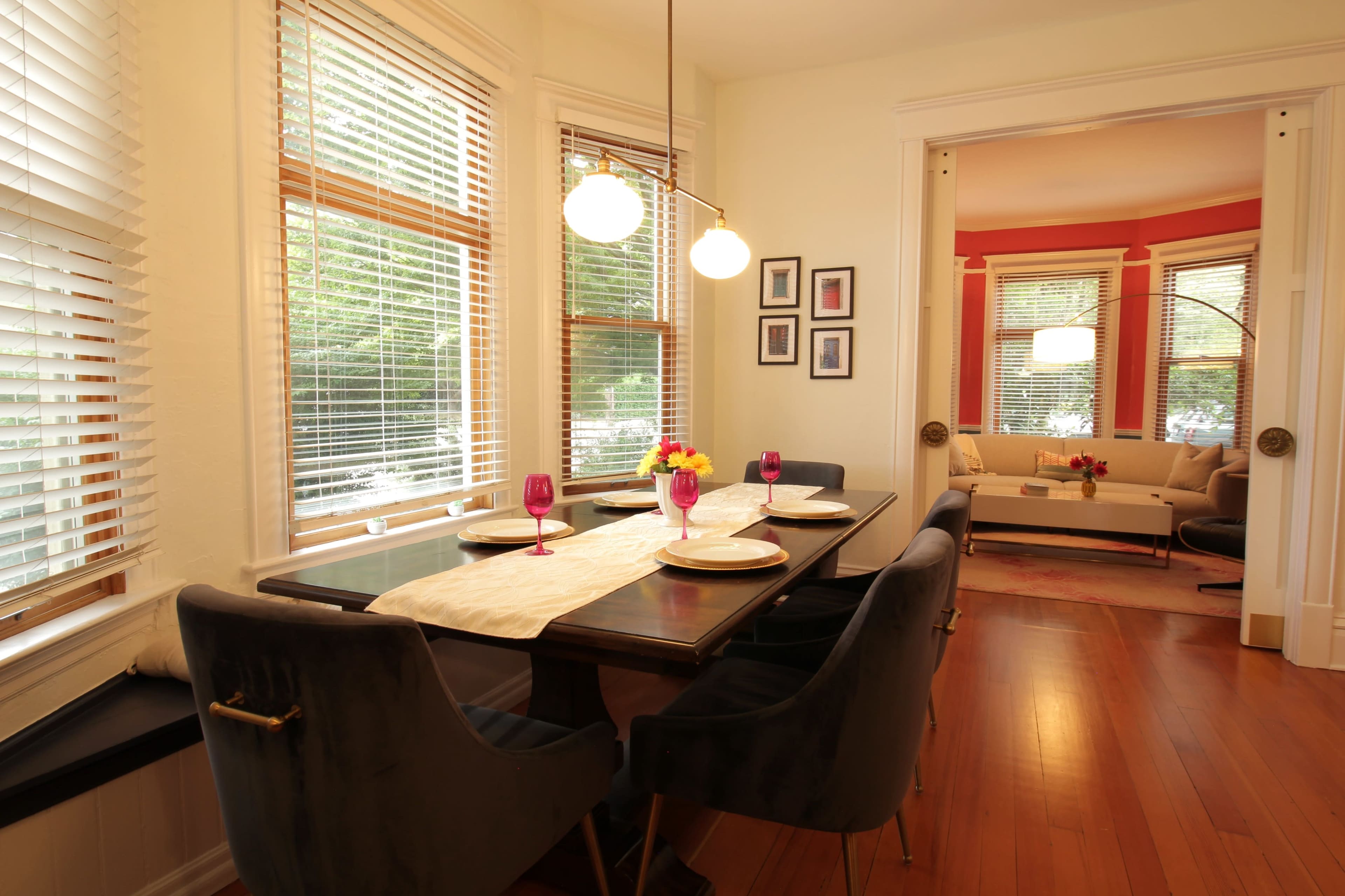 The image shows a dining area with a dark wooden table set for four, featuring plush chairs, pink glassware, and natural light streaming in through multiple windows.