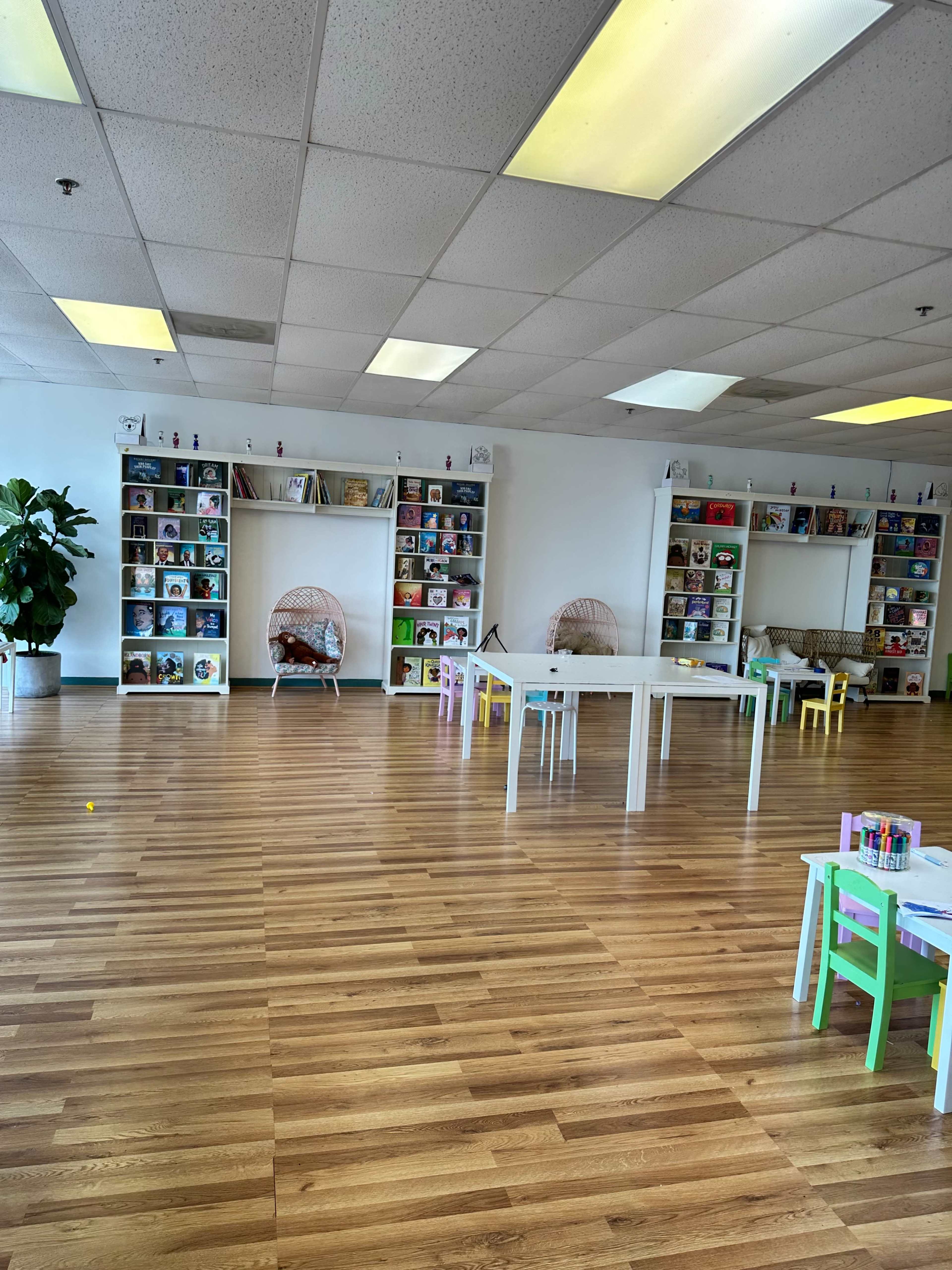 An empty children's classroom with bookshelves filled with books, two hanging chairs, and several small tables and chairs arranged for activities.