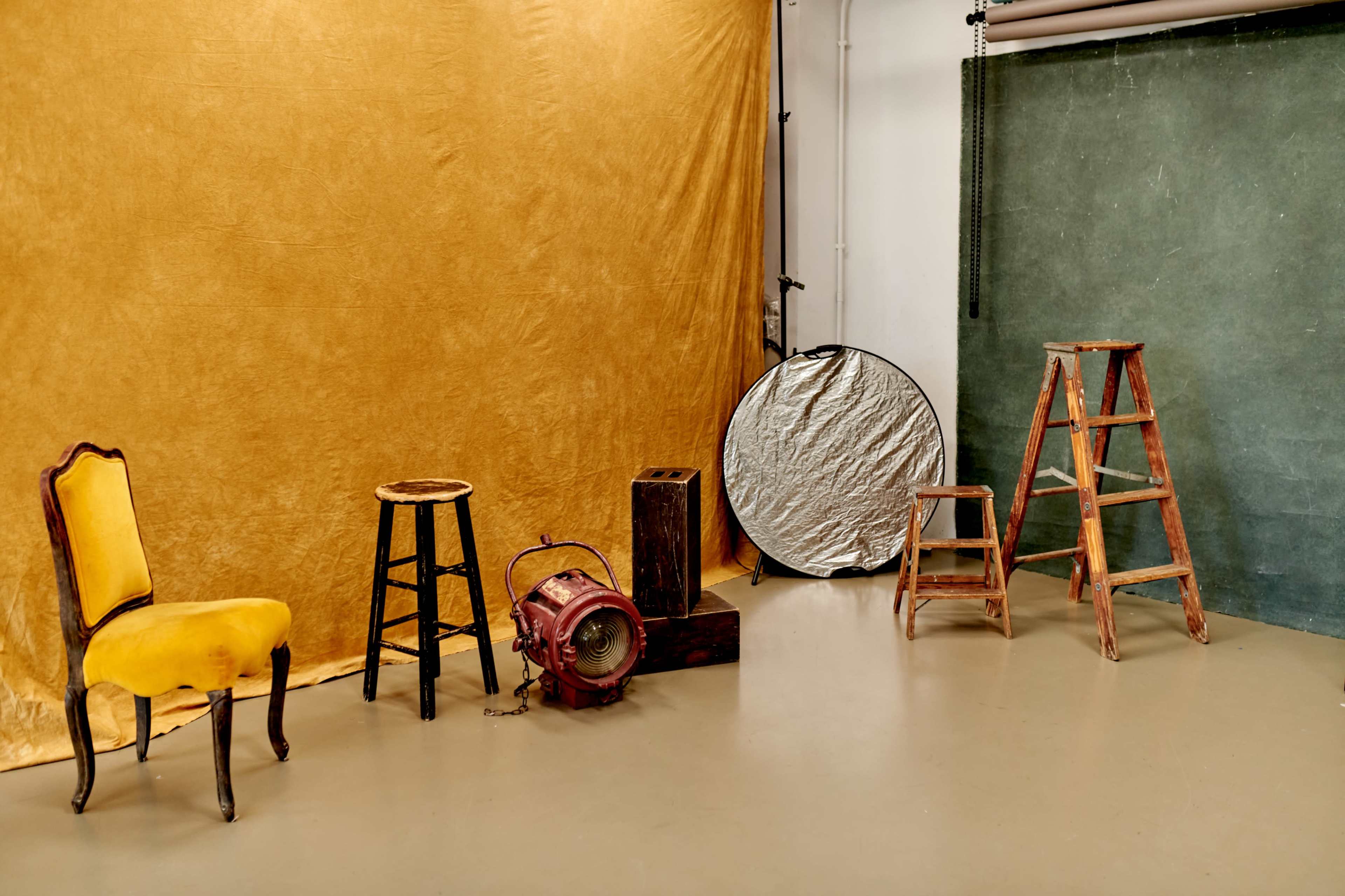 A photography studio setup with various props including a yellow chair, stools, a wooden ladder, and a reflector against a textured backdrop.