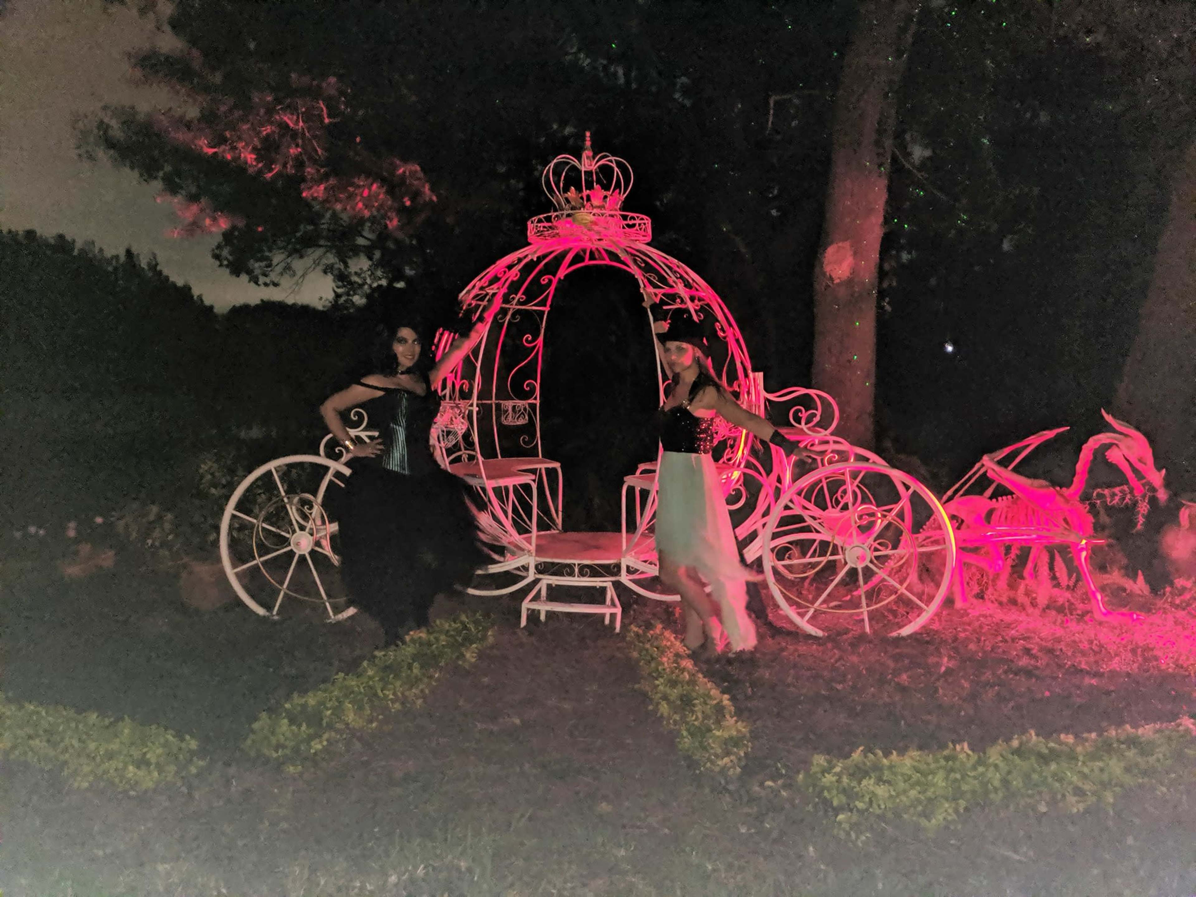 Two women pose beside a decorative, illuminated carriage in a garden at night.
