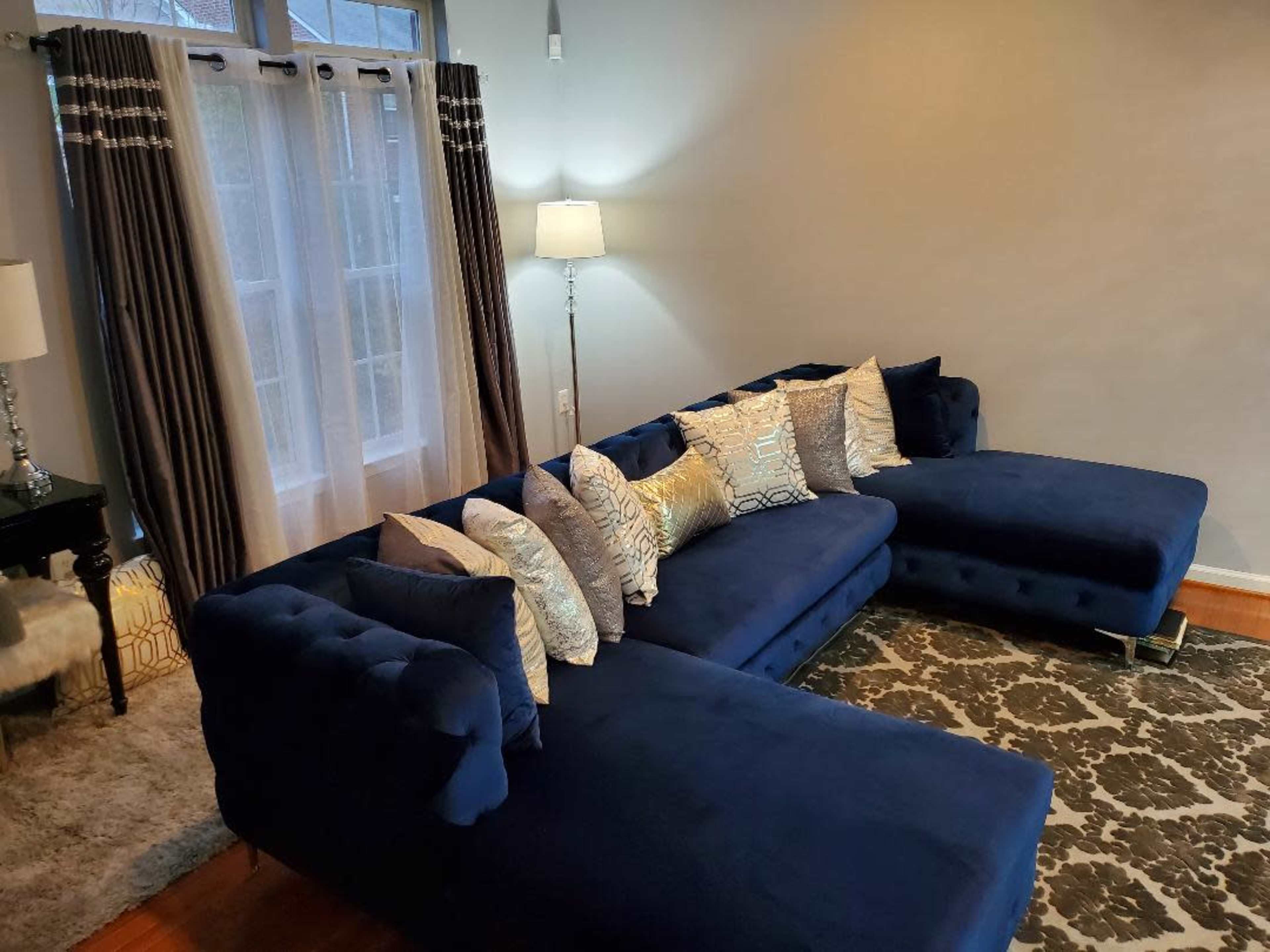 A modern living room features a navy blue sectional sofa adorned with decorative pillows, alongside a floor lamp and large windows draped with curtains.