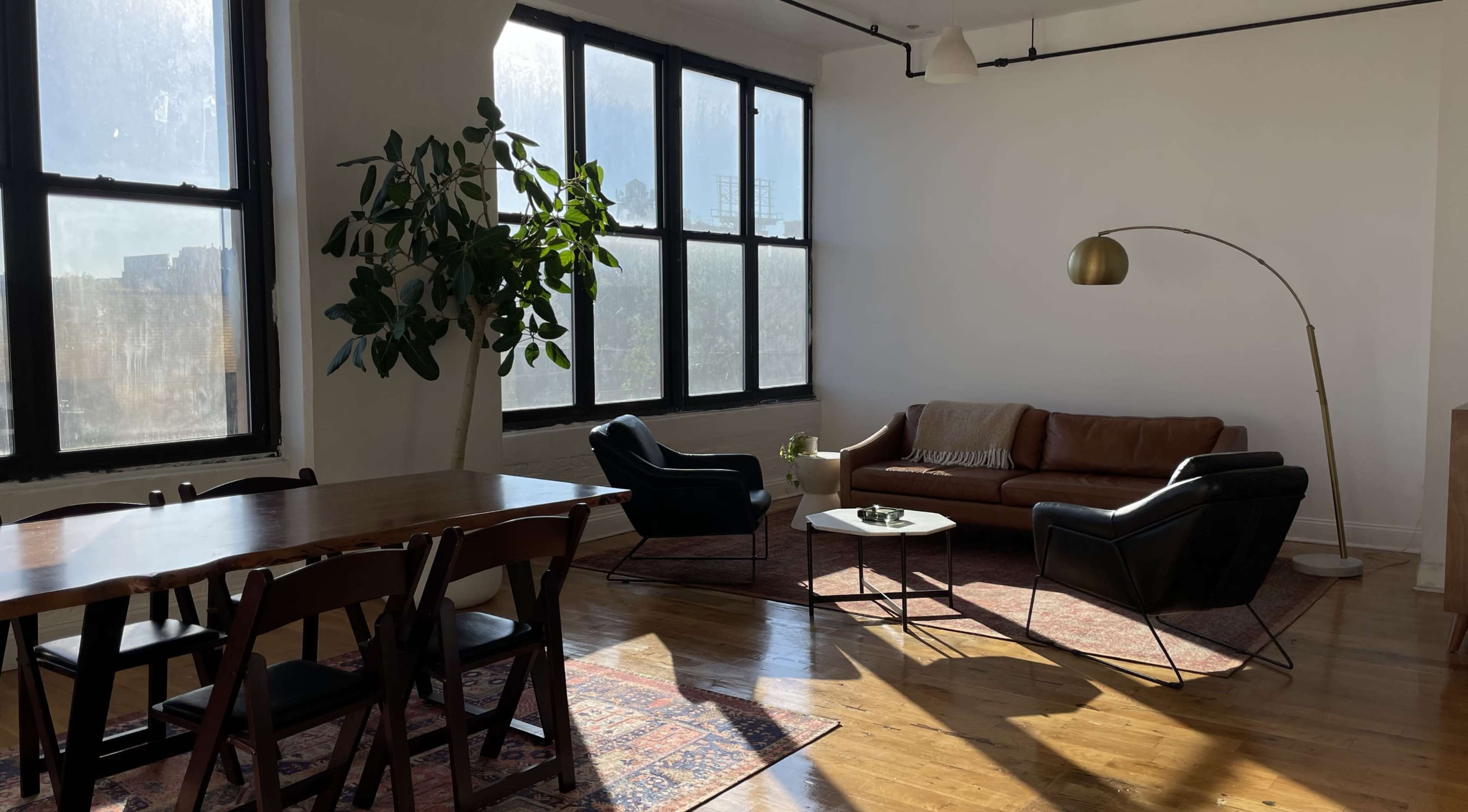 The image shows a bright, modern living space with large windows, a leather sofa, two black chairs, a dining table, and a potted plant.