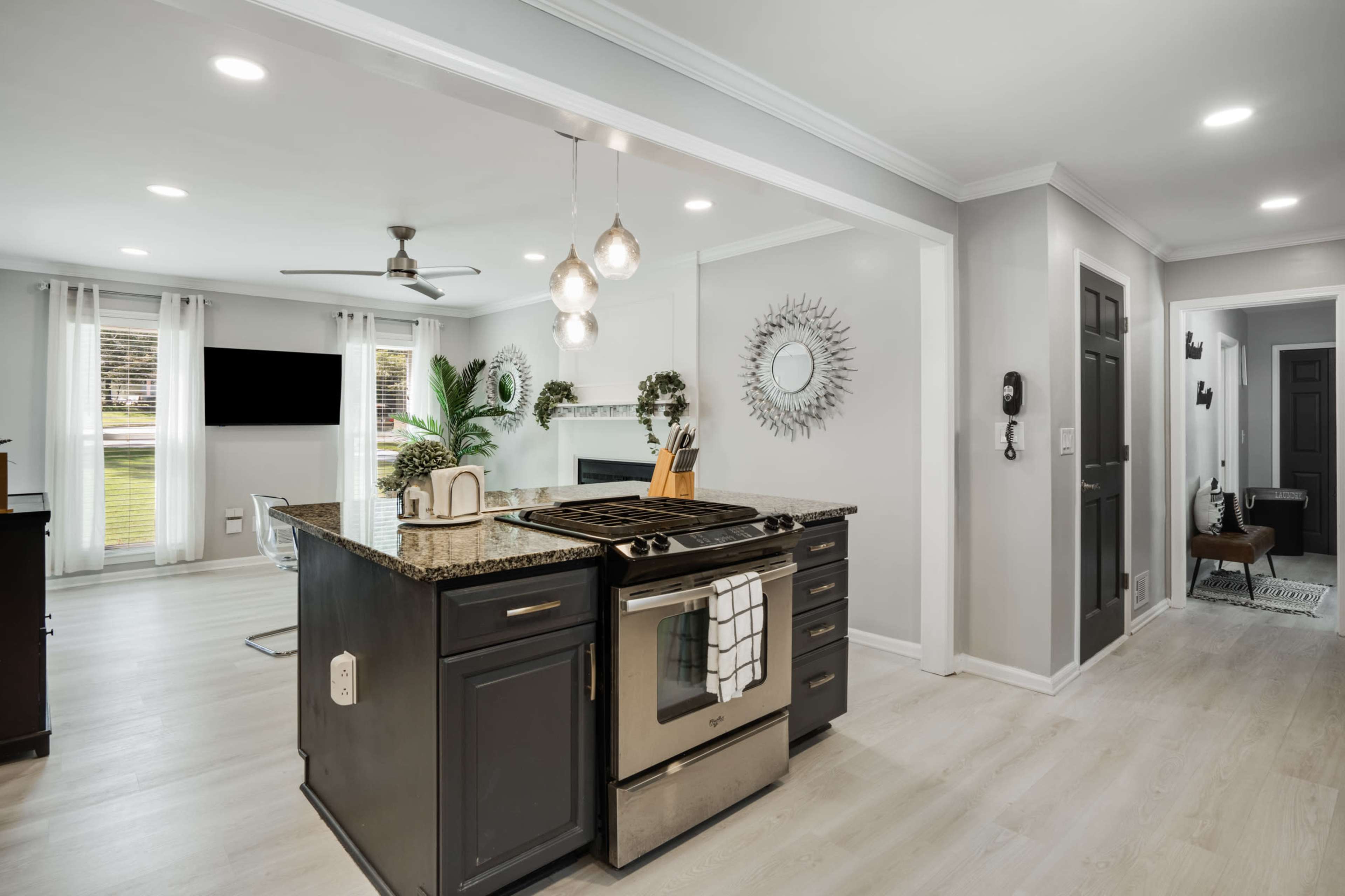 A modern kitchen with a central island, featuring a stove, granite countertops, and a view of an adjacent living area with natural light.