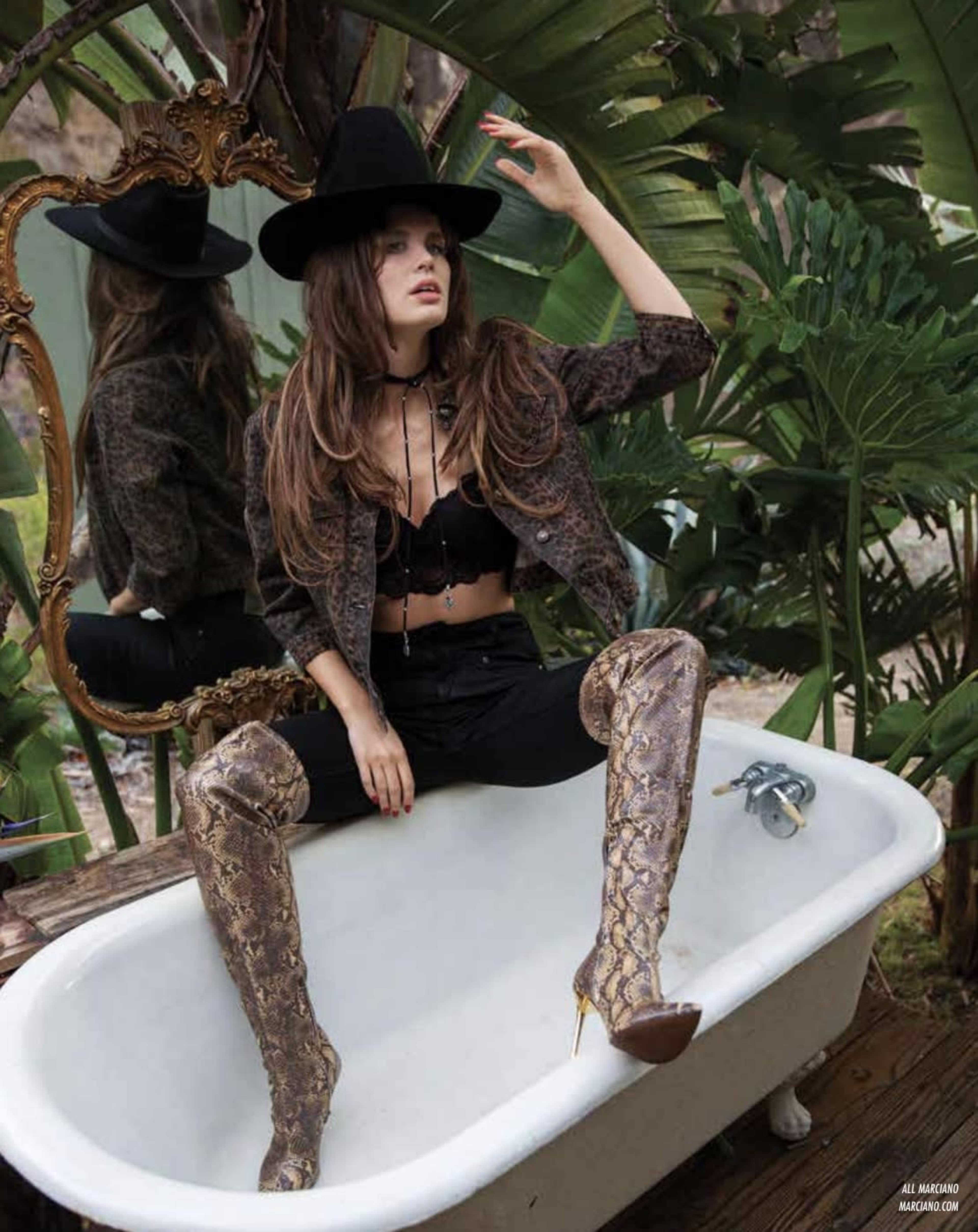A woman in a black crop top and patterned jacket poses in an outdoor setting, sitting in a vintage bathtub surrounded by plants, while wearing tall snake-print boots and a wide-brimmed hat.