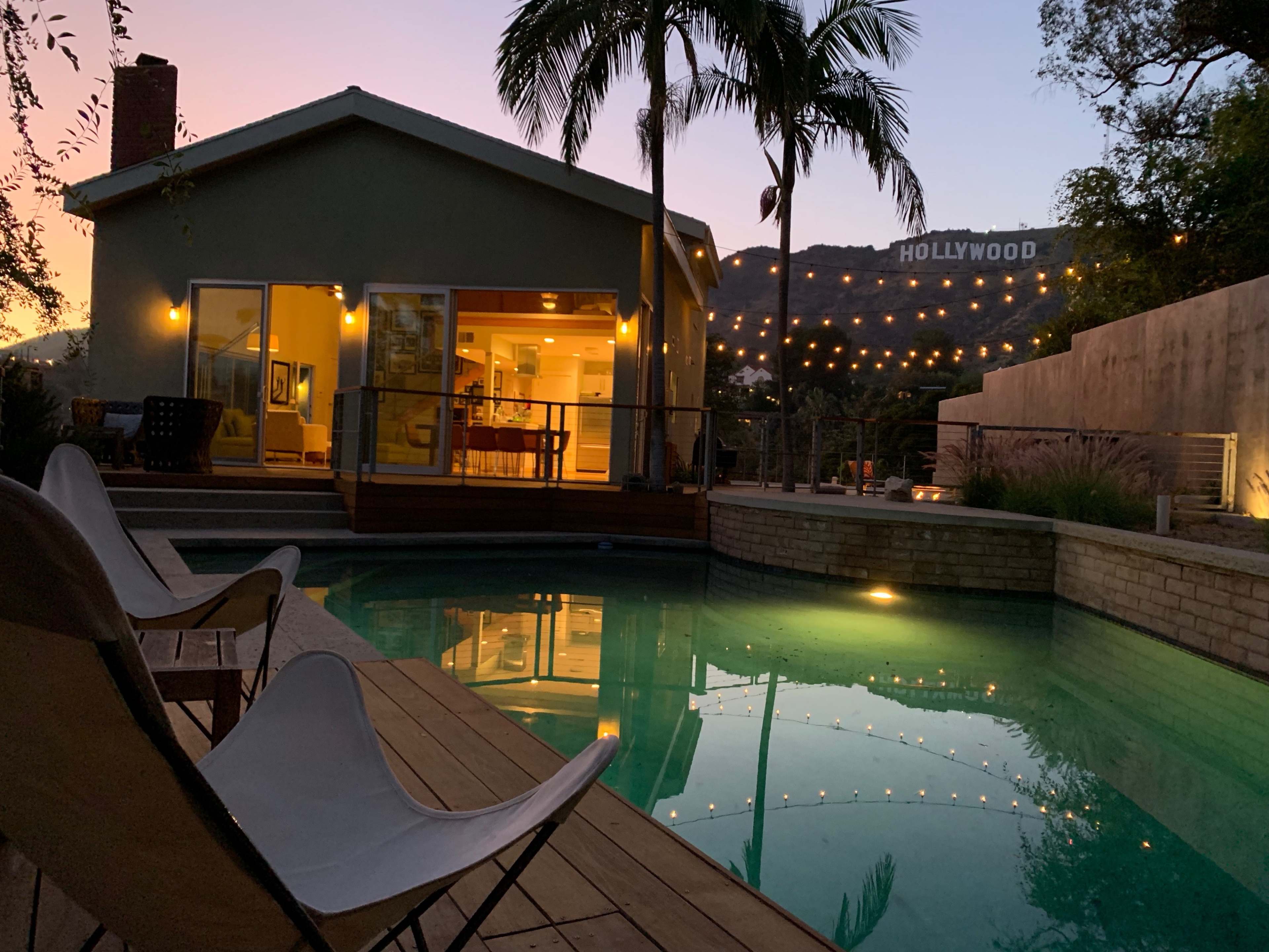 A house with large windows sits next to a swimming pool, reflecting the Hollywood sign in the background under a twilight sky.
