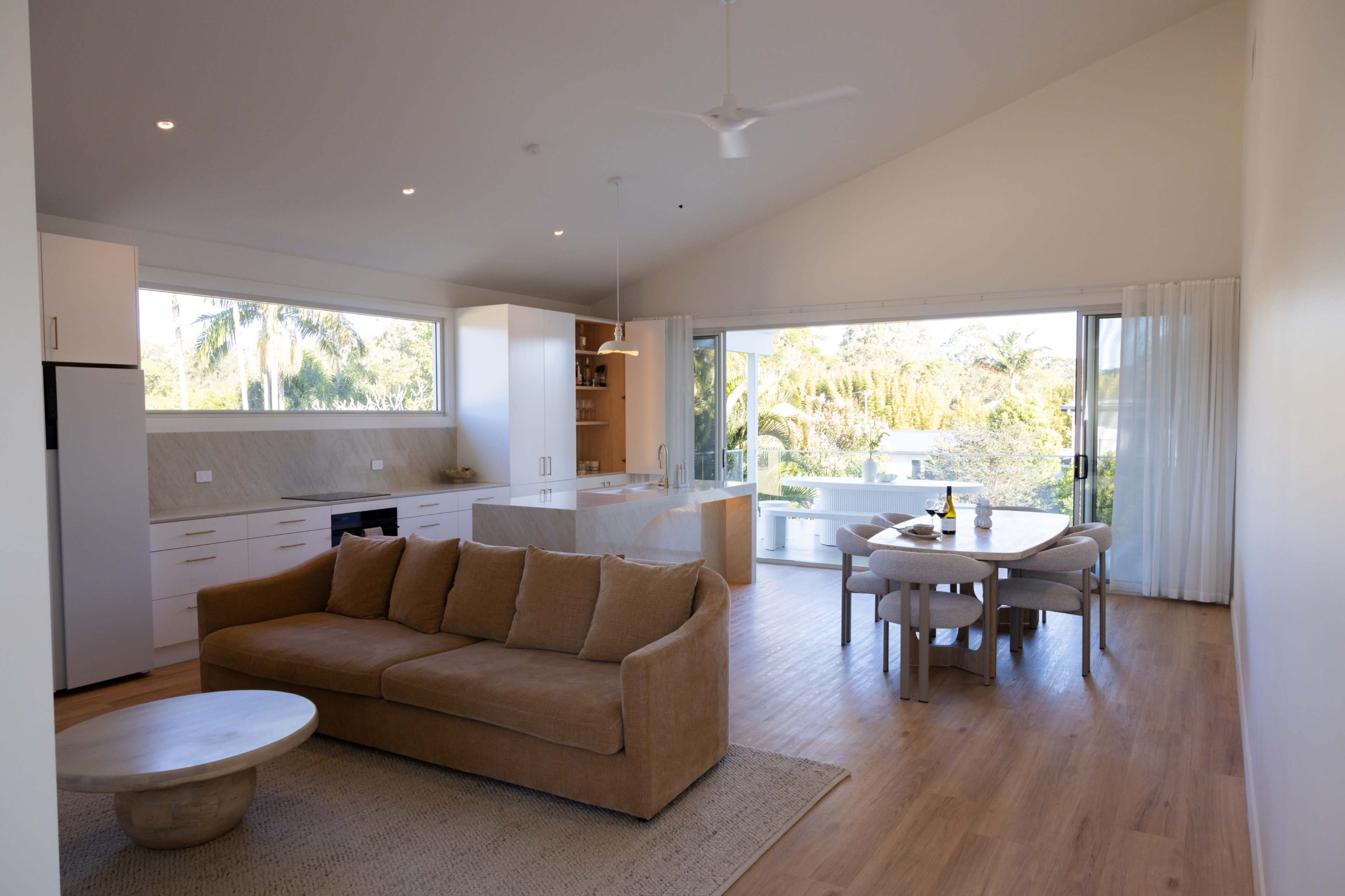 The image shows a modern, open-concept living area featuring a sofa, a wooden dining table, and large windows that offer views of the outside greenery.