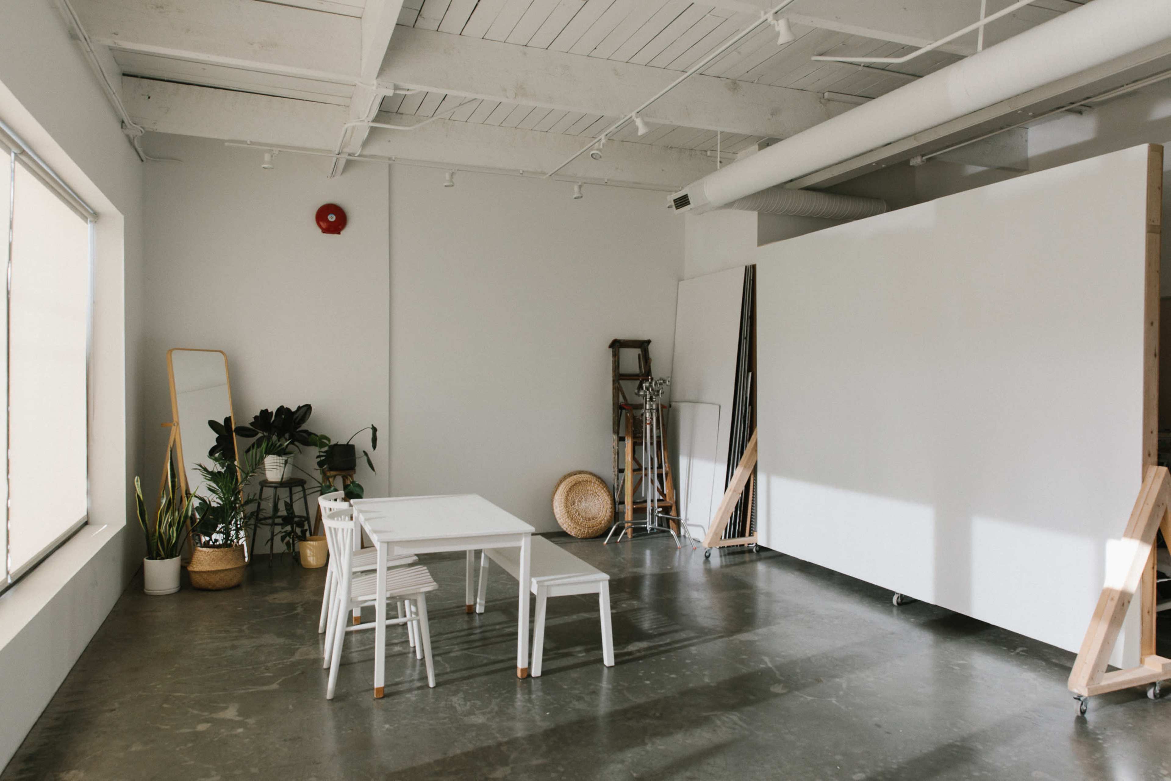 A minimalist studio space features a white table and chairs, a large white partition, potted plants, and various props against a concrete floor.